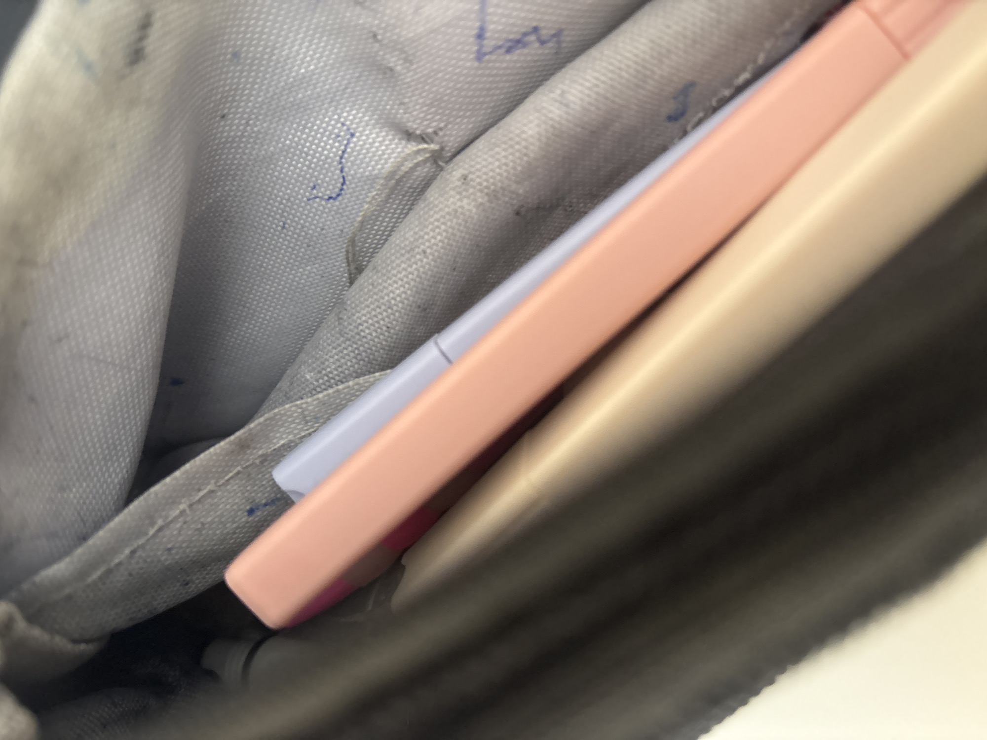 A close-up, indoor, eye-level shot depicts the interior of a dark gray pencil case. Inside the pencil case, three writing utensils are visible. The uppermost utensil is a light purple marker, partially obscured by a pink highlighter positioned directly in front of it. Beneath the pink highlighter is a beige-colored pencil. The fabric lining of the pencil case is light gray and shows signs of wear and smudging, with some blue ink marks visible. The lighting is artificial and subdued, casting shadows that contribute to the overall dark tone of the image. There are no people or external environmental elements visible. The scene is devoid of any contextual clues to determine its precise location within the city of Georgiyevsk, Russia, or any other specific geographical area.