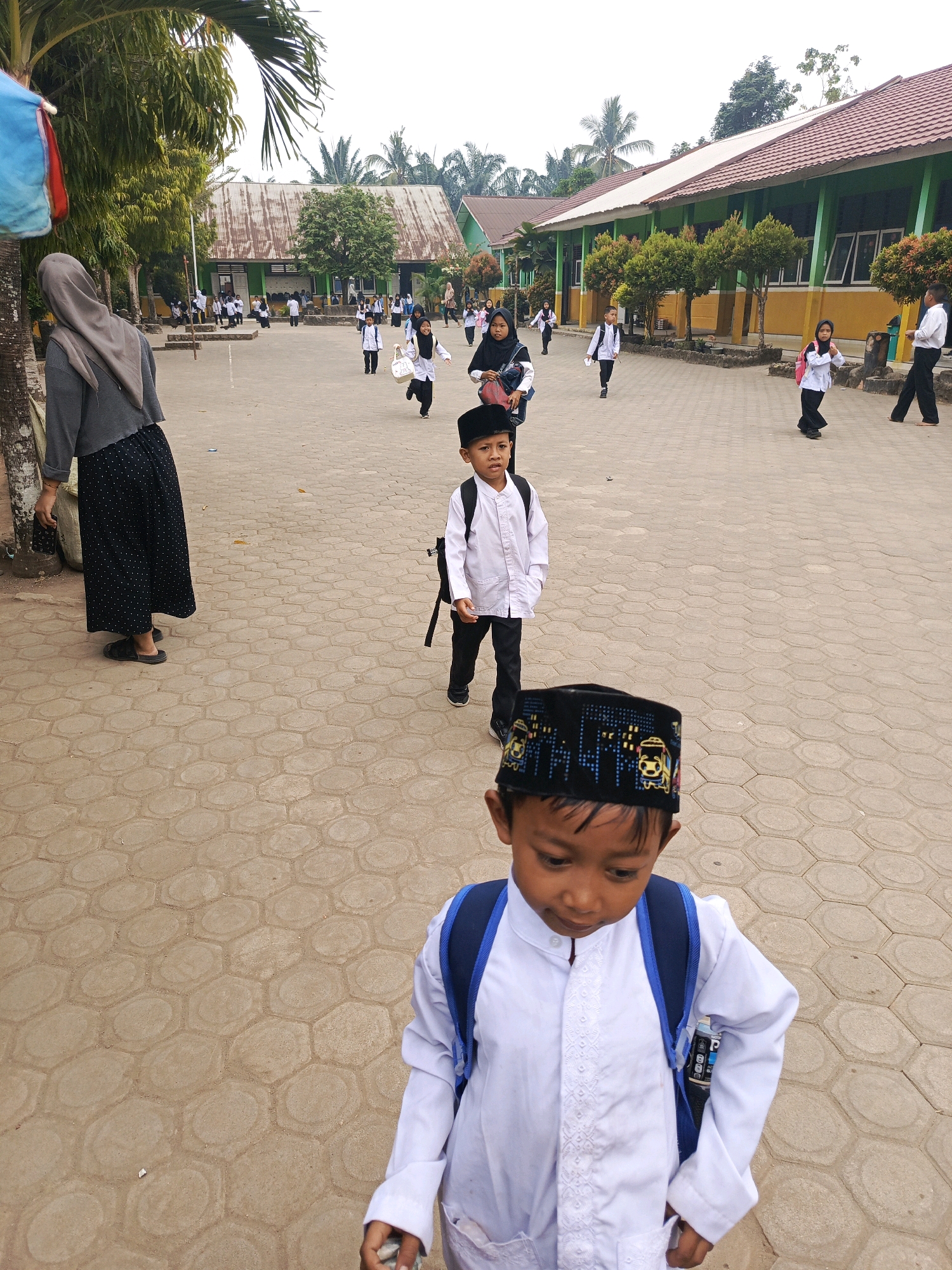 In the city of Megang Sakti II, Indonesia, a group of children in school uniforms are walking across a paved courtyard in front of a school building. The school building has green walls and a red roof, with trees and foliage in front of it. The foreground features a young boy wearing a white shirt, black pants, and a black and yellow patterned cap, with a blue backpack. Behind him, another boy in a white shirt and black pants with a black backpack walks away from the camera. Other children, some carrying backpacks, are scattered throughout the courtyard, walking towards or away from the school. The ground is paved with hexagonal interlocking tiles. A woman in a grey hijab and a black and white patterned skirt is visible on the left side of the image, standing near a tree. The sky is overcast.