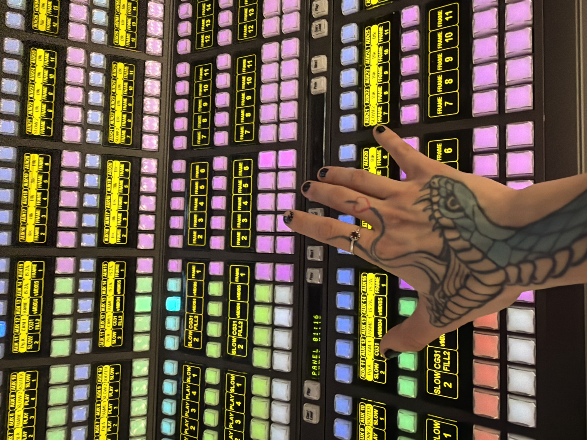 A medium shot depicts a portion of a large control panel, likely for broadcast or media production, being operated by a person. The panel is black with numerous illuminated buttons and labels. The buttons are arranged in rows and columns, with various colors and text indicating their functions. The panel's labels include "FRAME," "AUX," "PLAY," "SLOW," and others suggesting control over video sources, transitions, and effects. The time on the PANEL reads "01:16."
A human hand with black nail polish and a snake tattoo is positioned over the panel, seemingly in the act of pressing or manipulating one of the buttons. The hand has a ring on the ring finger and a small heart tattoo on the inner wrist.
Given the nature of the control panel, the environment is likely a television studio, broadcast facility, or a similar space dedicated to media production. There is no information available to determine the location in the city of Sofia, Bulgaria.