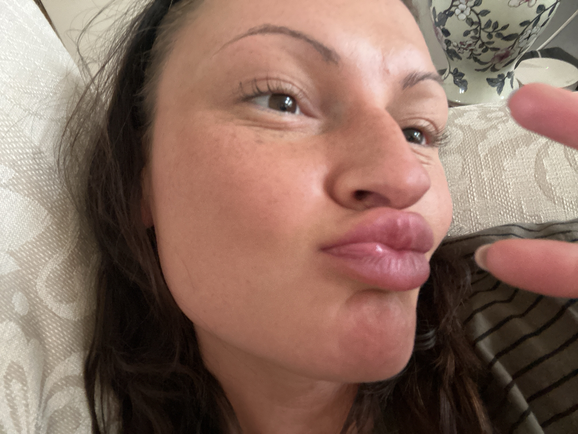 A close-up selfie of a woman with brown hair, light skin, and brown eyes. She is making a kissing face with pursed lips. A finger is partially visible in the lower right corner. The background includes a patterned white surface, possibly a cushion or upholstery, and a floral patterned vase. The location is undetermined; there's no clear evidence indicating Tunbridge Wells, United Kingdom.
