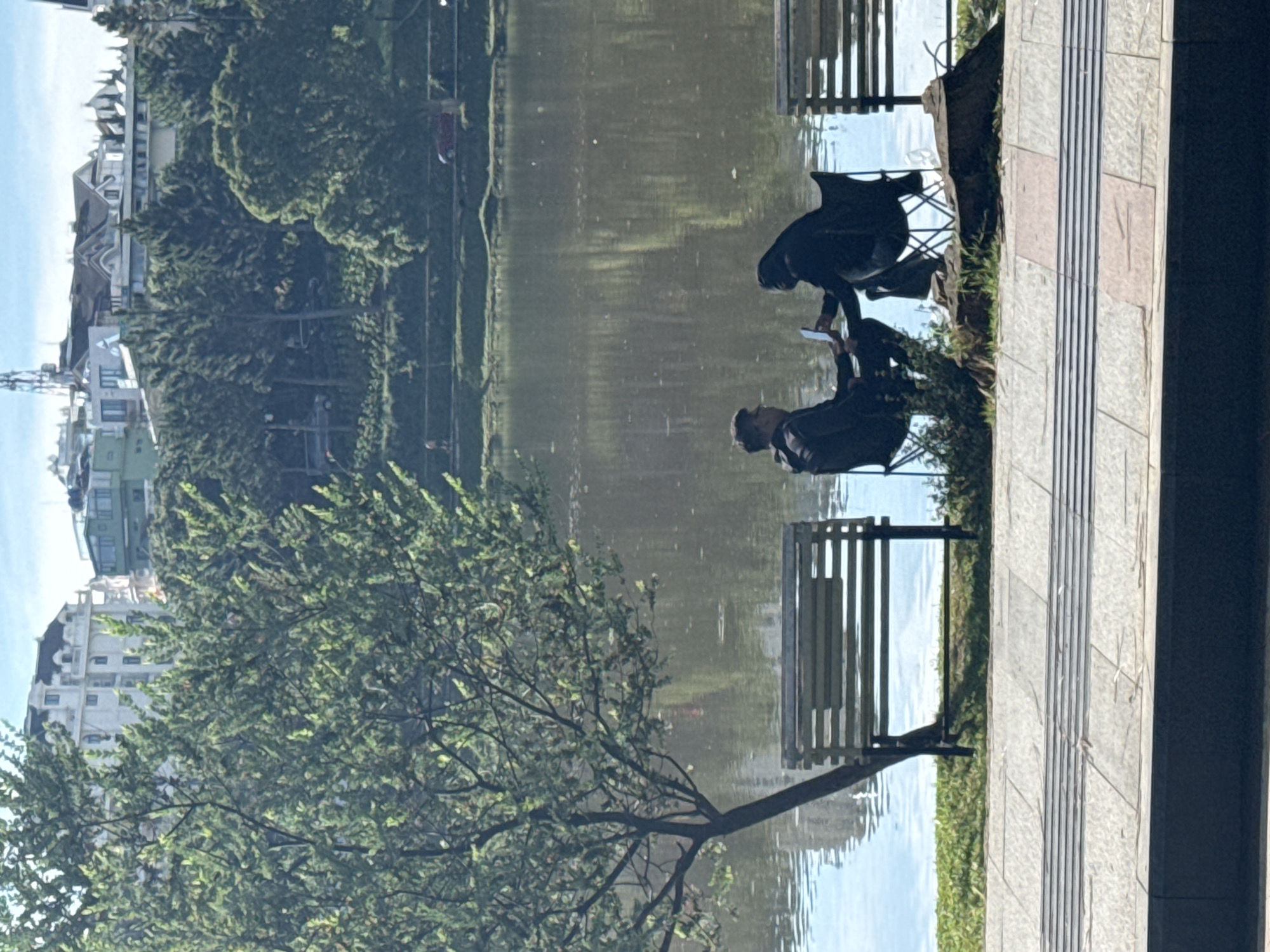 The image shows a lake in Da Lat, Vietnam. On the bank of the lake, there is a stone pathway with two benches. Two people are sitting on one of the benches, and one of them is holding a piece of paper. A tree with green leaves is on the left side of the image. In the background, there are buildings and trees. The water in the lake reflects the buildings and trees.