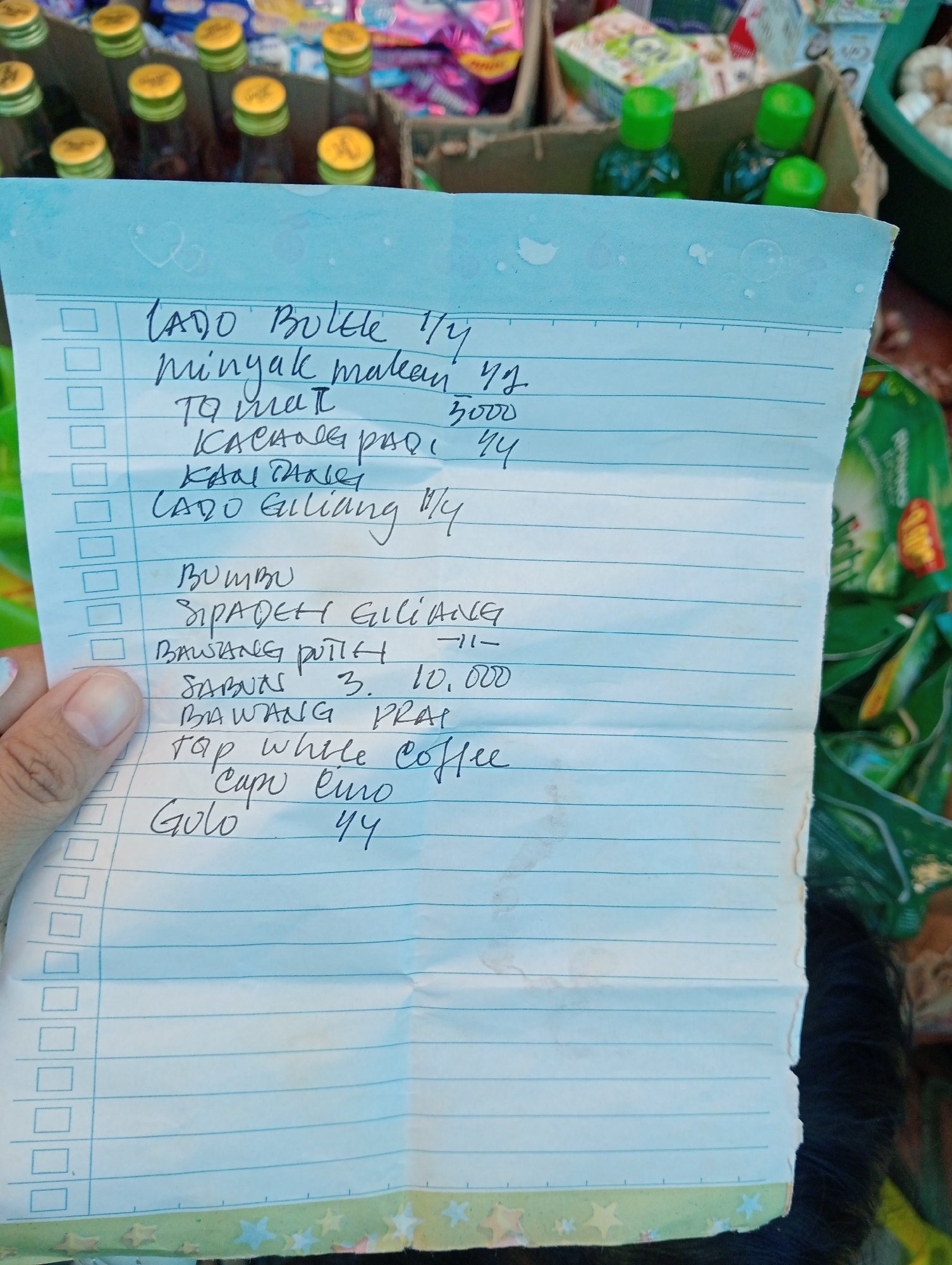A handwritten grocery list is held in the foreground, obscuring a display of bottled goods and packaged items in the background. The list, written in blue ink on lined paper, details various items and quantities. In the background, several glass bottles with yellow caps and green lids are visible, along with cardboard boxes containing other products. The setting appears to be a local market or shop.