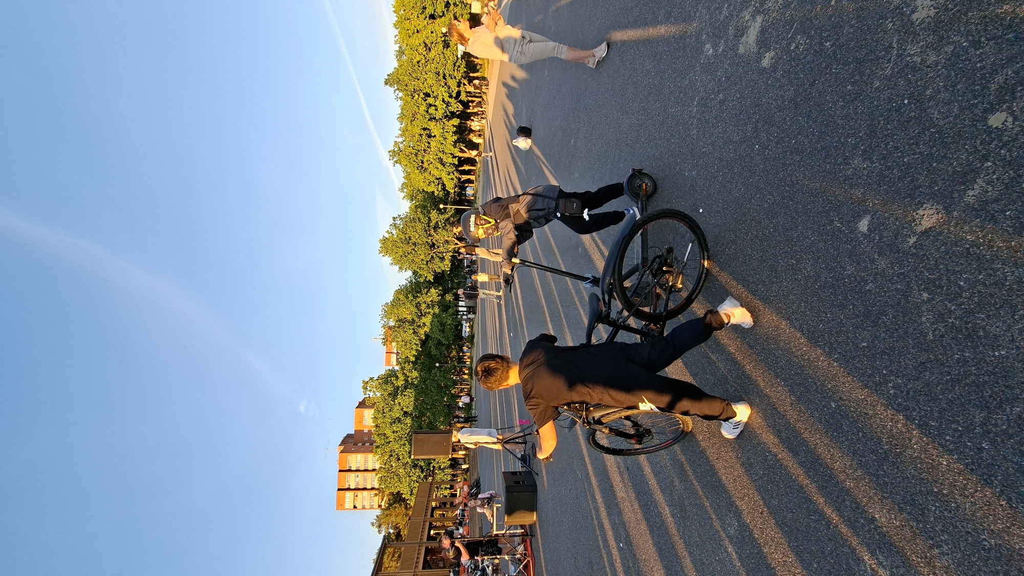 In a public plaza in Tallinn, Estonia, two young individuals are engaged in an interaction involving a bicycle and an electric scooter. A boy in a black tracksuit and white sneakers is positioned behind a black bicycle, with his left hand on the handlebars, appearing to be holding it stationary. To his right, another individual, wearing a grey jacket, black pants, and a helmet with protective goggles, is standing on an electric scooter. The scooter rider's right hand is on the handlebars, and their left hand is held out, possibly in an attempt to take or receive something from the boy with the bicycle. The background features a clear blue sky with faint contrails, a line of green trees, and several buildings. In the mid-ground, there's a stage setup with speakers and a camera, suggesting an event is taking place or has recently concluded. Other people are visible in the distance, some seated and others standing, enjoying the outdoor setting. The ground is paved, and long shadows from the trees and people stretch across it, indicating the low angle of the sun, likely during the late afternoon or early evening.