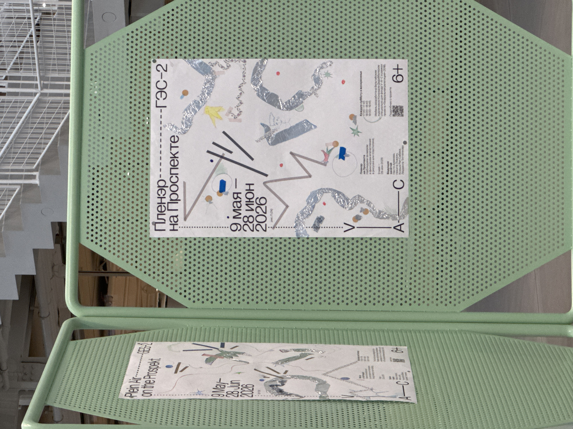 The image shows two posters displayed on green, perforated, hexagonal metal frames. Both posters are about an event called "Plenair on the Prospekt" or "Пленэр на Проспекте".
The poster on the left is partially visible and reads "Plein Air ---GES-2 on the Prospekt" and lists dates "9 May—28 Jun 2026".
The poster on the right is fully visible and in Russian. It has the title "Пленэр на Проспекте -----ГЭС-2". It also lists dates "9 мая — 28 июня 2026". Below this, there is information about the event, including "Каждую субботу и воскресенье" (Every Saturday and Sunday) with times "17:00–17:45" and "19:00–19:45". It mentions "Мебель разработана бельгийским художником Конрадом Дюдоблером..." (Furniture designed by Belgian artist Conrad Dudobler...). There is also a section for "Подробнее о проекте" (More about the project) with a QR code, and the age rating "6+". Further down, it lists "Ведущие занятия" (Leading classes) with names including "Настя Артеева", "Никита Ложкин", "Ксюша Реп Сабака", and "Людмила Чен-чегова".
The posters are displayed outdoors in an urban environment. The background shows a concrete structure with stairs and railings, suggesting it might be part of a public space or an exhibition area. The overall color scheme is dominated by the mint green of the display frames and the white of the posters, with graphic elements in various colors and metallic silver foil. The image does not provide specific information about the location in Moscow, Russia, but the presence of Cyrillic text on the poster and the style of the urban background are consistent with a Russian setting.