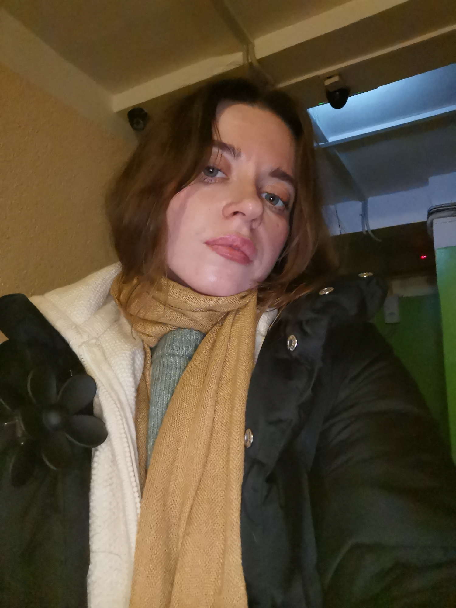 The image captures an adult woman, likely taking a selfie, in what appears to be an indoor, possibly commercial or public setting. She has shoulder-length brown hair, blue-green eyes, and light makeup, with a serious or slightly pouting expression.
She is dressed for cold weather, wearing a white or off-white textured jacket over a light blue-green knitted top, and a camel-colored scarf wrapped around her neck. A black puffer jacket with silver snaps is visible on her right side, and her left hand is adorned with a black glove featuring a flower-like embellishment.
The background suggests an institutional or industrial environment. To her left is a beige, textured wall. To her right, a bright green wall is visible, along with what appear to be pipes or conduits near the ceiling. Two dark-colored security cameras are mounted on the ceiling or high on the wall in the background, one above her head on the left and another further back on the right, indicating a monitored space. Faint red indicator lights are also visible on the right side. The lighting is artificial and casts a cool tone in some areas. There is no visible text in the image. The exact time of day or specific location is not discernible, but the attire suggests a cooler climate.