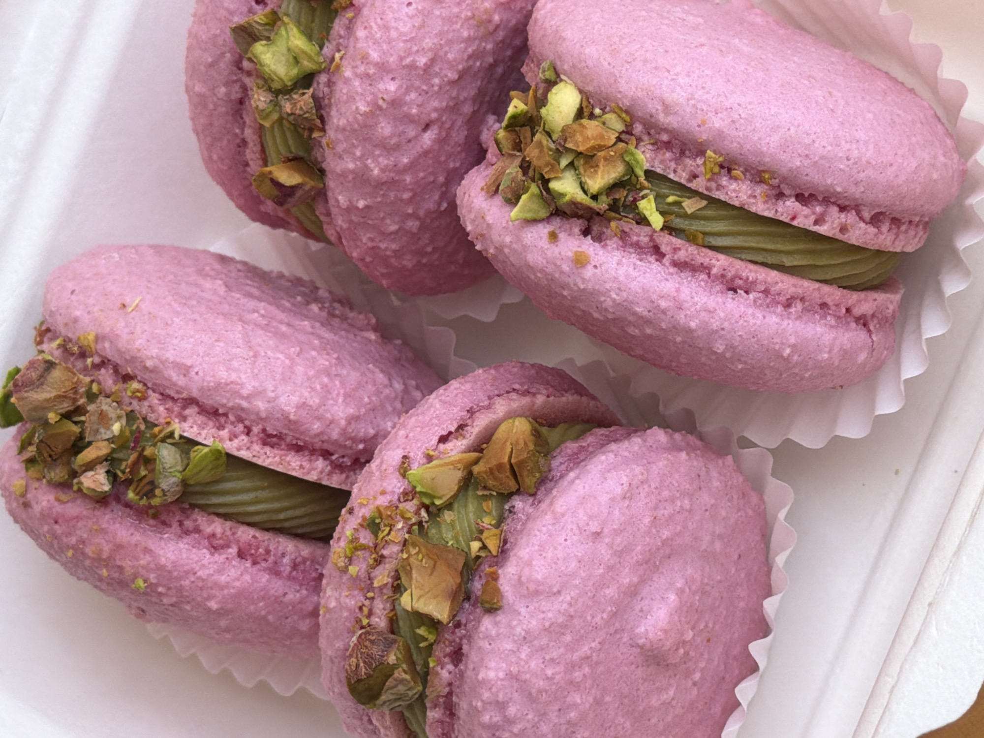 The image shows a close-up of four pink macarons with green filling and chopped pistachios, arranged in white cupcake liners and placed in a white container. The scene is likely indoors due to the controlled lighting and the packaging of the macarons. While the city of Tel Aviv, Israel, is specified, there is no information in the image itself to directly confirm the location. Therefore, the description focuses on the visible elements of the image only.