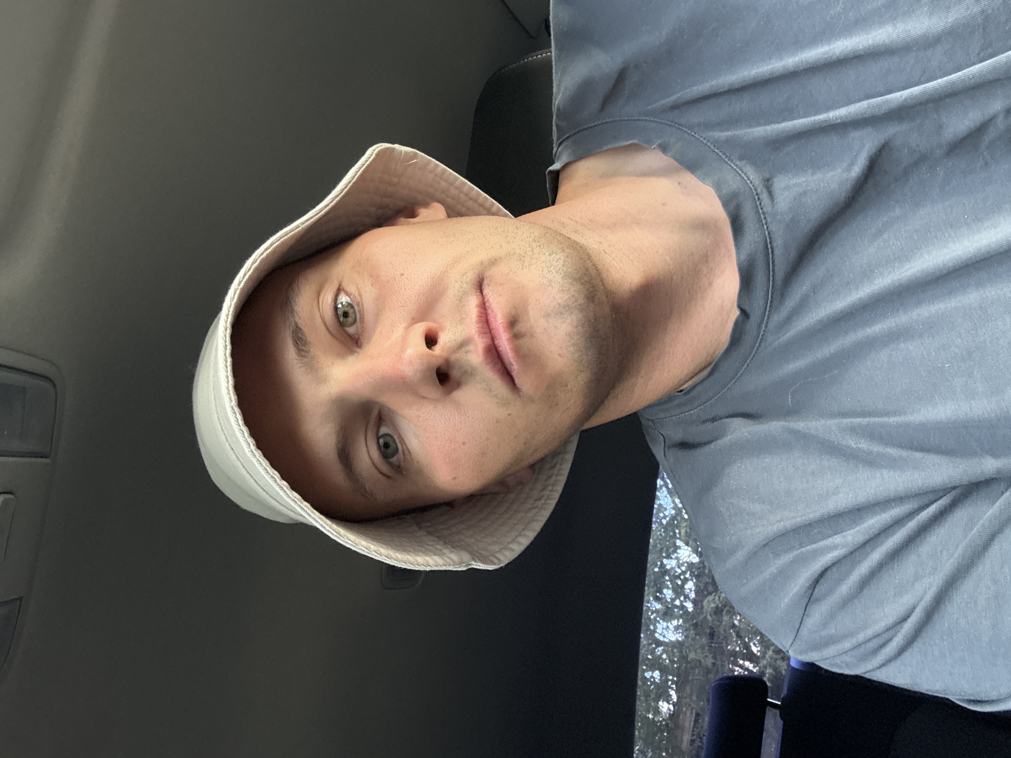 A man wearing a white bucket hat and a grey t-shirt takes a selfie in a car. The man's face is partially visible, with his eyes looking towards the camera. The interior of the car is dark, with the ceiling and dashboard visible. Outside the car window, trees and foliage can be seen. The image was taken in Chaikovsky, Russia.