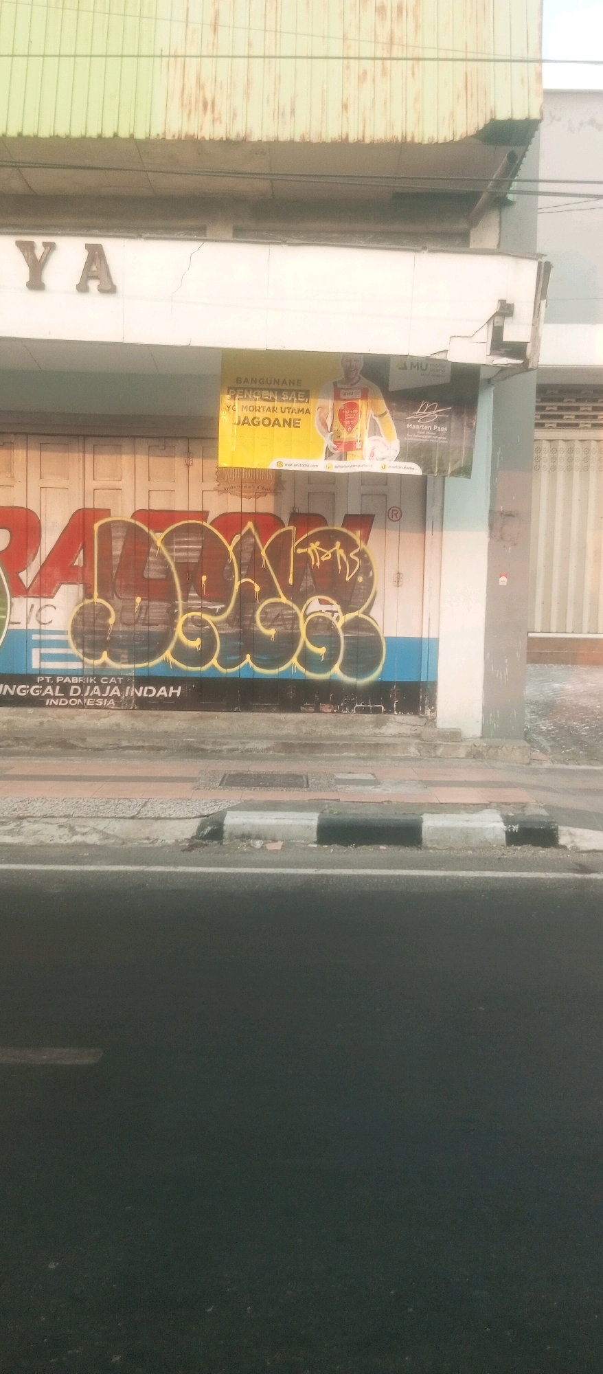 The image captures a storefront in Kediri City, Indonesia, adorned with graffiti. The storefront is likely part of a commercial building, with a large green, corrugated metal awning extending over the upper section. Below the awning, a white facade features signage that reads "YA". A yellow banner advertising "BANGUNANE PENGENC SA", "YO MORTAR UTAMA JAGOANE", and an image of a man holding a soccer ball is affixed to the wall. The main feature of the lower portion of the storefront is a large graffiti tag in brown with a yellow outline, spray-painted over a blue and white striped background. Text beneath the graffiti reads "PT. PABRIK CAT" and "UNGGAL DJAJA INDAH INDONESIA". The street in front of the building has a sidewalk with a black and white striped curb, and a paved road. The lighting suggests it may be early morning or late afternoon.