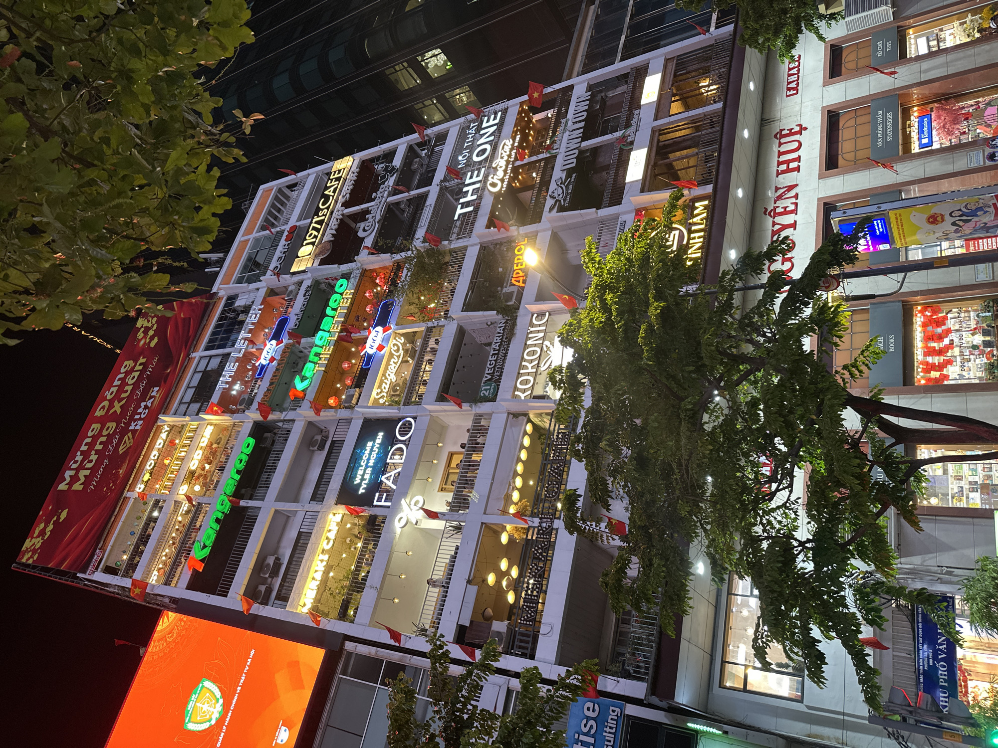 The image shows a multi-story building in Thủ Đức, Vietnam, at night. The building's facade features numerous businesses with brightly lit signs, including restaurants, cafes, and shops. Each business has its own sign and decorative elements. Red Vietnamese flags are visible along the building's edge. Banners with Vietnamese text are draped on the building's left side. Below the building, there's an LED screen and other store signs. Trees partially obscure the view of the building and the street.