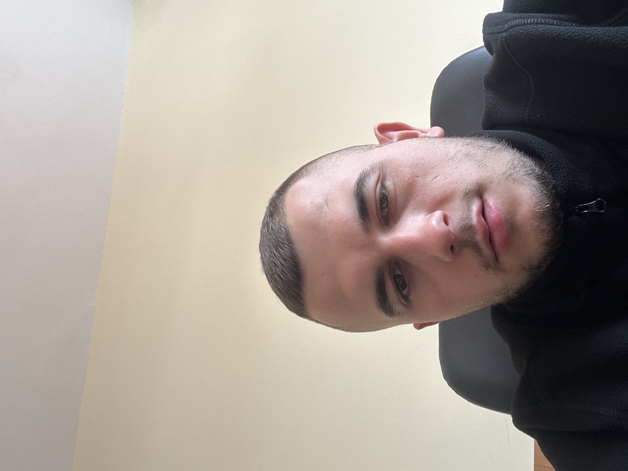 A close-up, slightly rotated image shows a man with a buzz cut haircut and light stubble. He is wearing a black fleece jacket and seated in a black chair. The background consists of a plain, light yellow or beige wall in the upper portion and a lighter gray wall in the bottom left corner. The image lacks identifying details to confirm a location in Plovdiv, Bulgaria. No specific actions or interactions are apparent.
