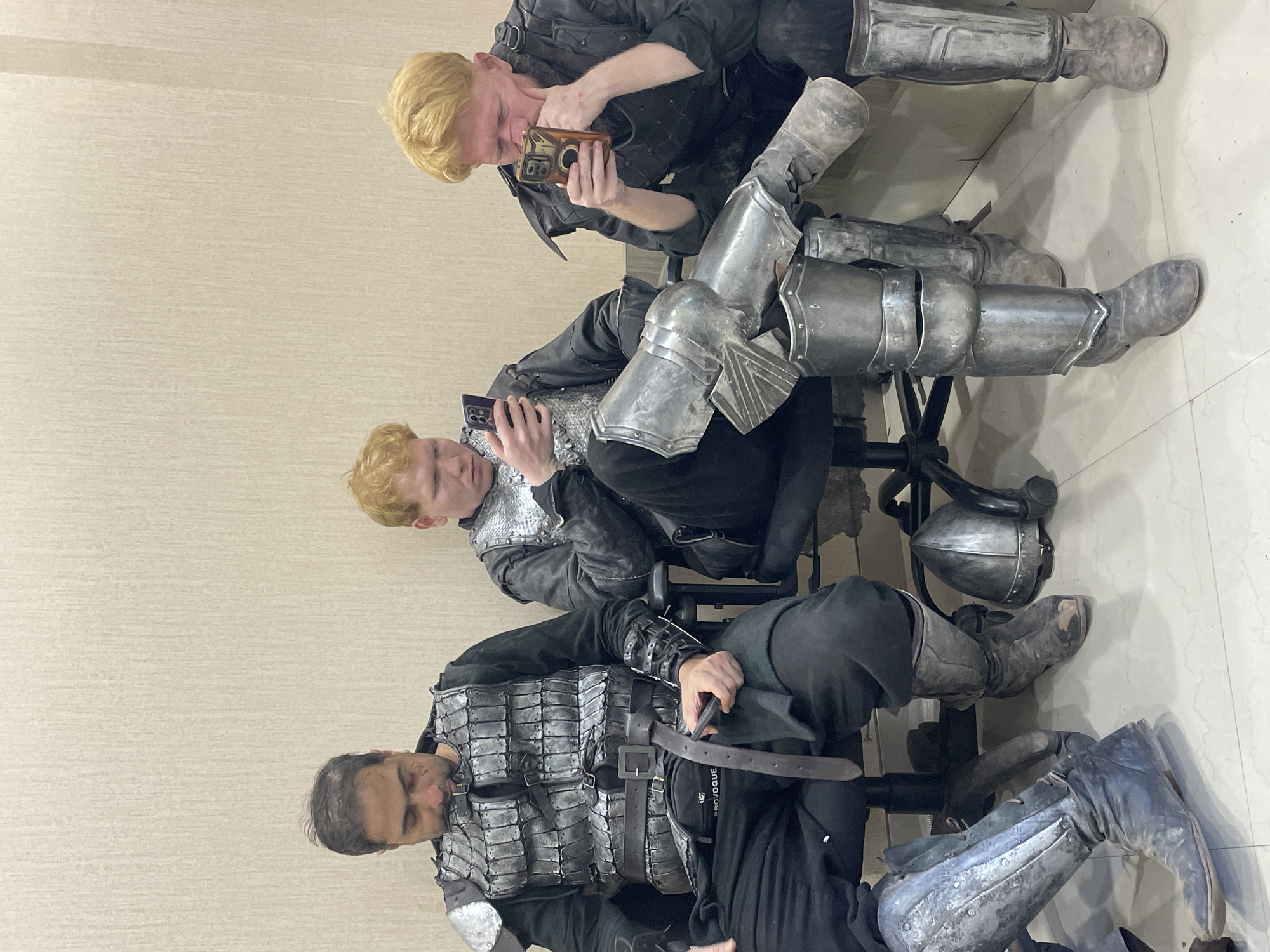 Three people are seated in chairs against a light beige wall with what appears to be a tile floor. All three are wearing what appears to be armor, including a metal breastplate, arm and leg coverings, and dark clothing underneath. The person at the top of the image has blonde hair and is looking at a phone. The person in the middle also has blonde hair and is looking at a phone as well. The person at the bottom of the image has dark hair and is holding what looks like a leather belt. A helmet is sitting on the floor beside the middle person. The location is not discernible from the image. There is no evidence to suggest that the location is in the city of Mumbai, India.