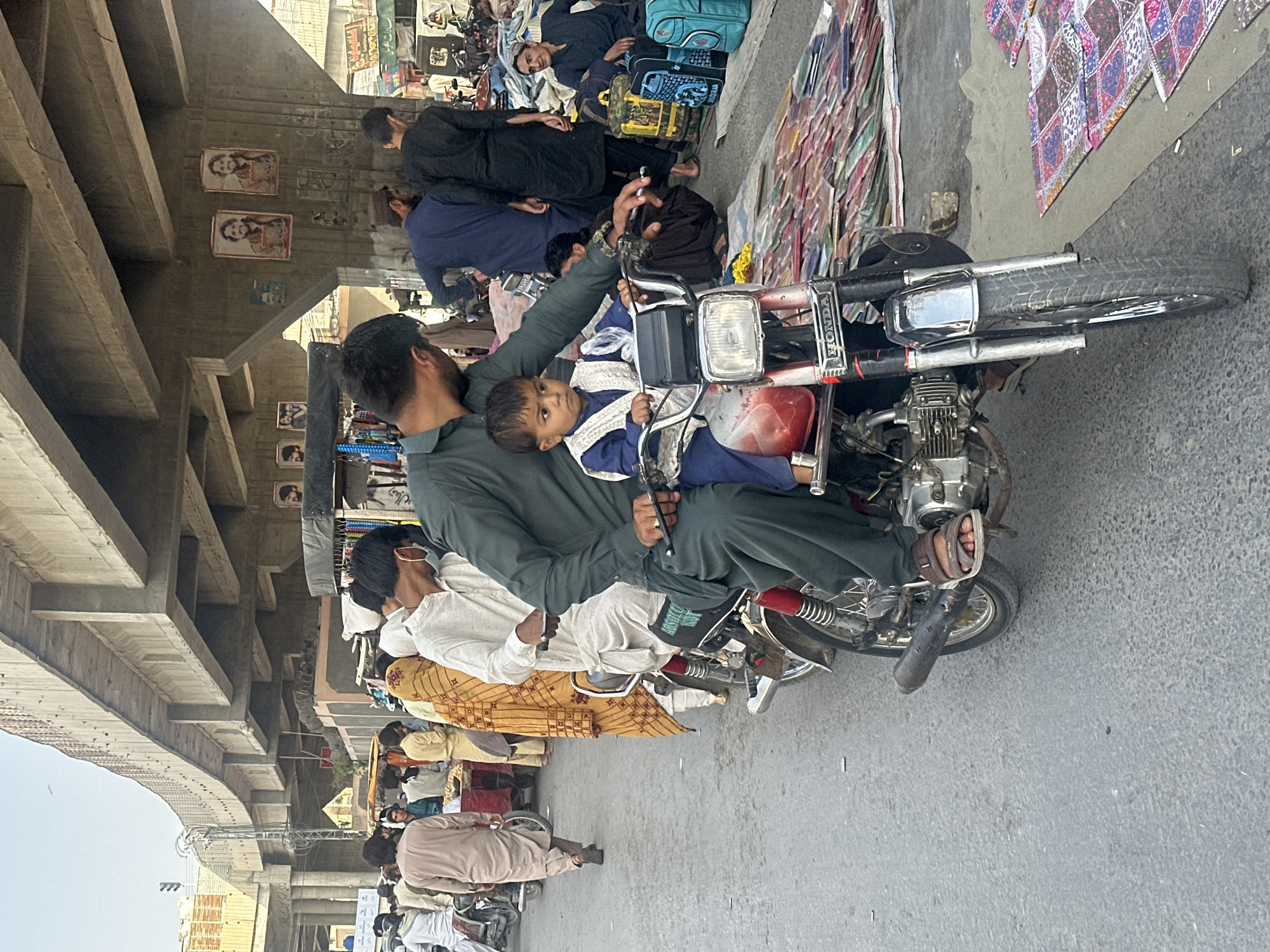 A street scene in Quetta City Tehsil, Pakistan. A man in a dark green shirt and trousers sits on a red Honda motorcycle, holding a small child dressed in blue. The motorcycle is parked on a paved road, close to a sidewalk. Several other people are nearby, some walking, others sitting. A pedestrian bridge is visible overhead, its concrete pillars adorned with pictures. A variety of goods are displayed on the sidewalk.