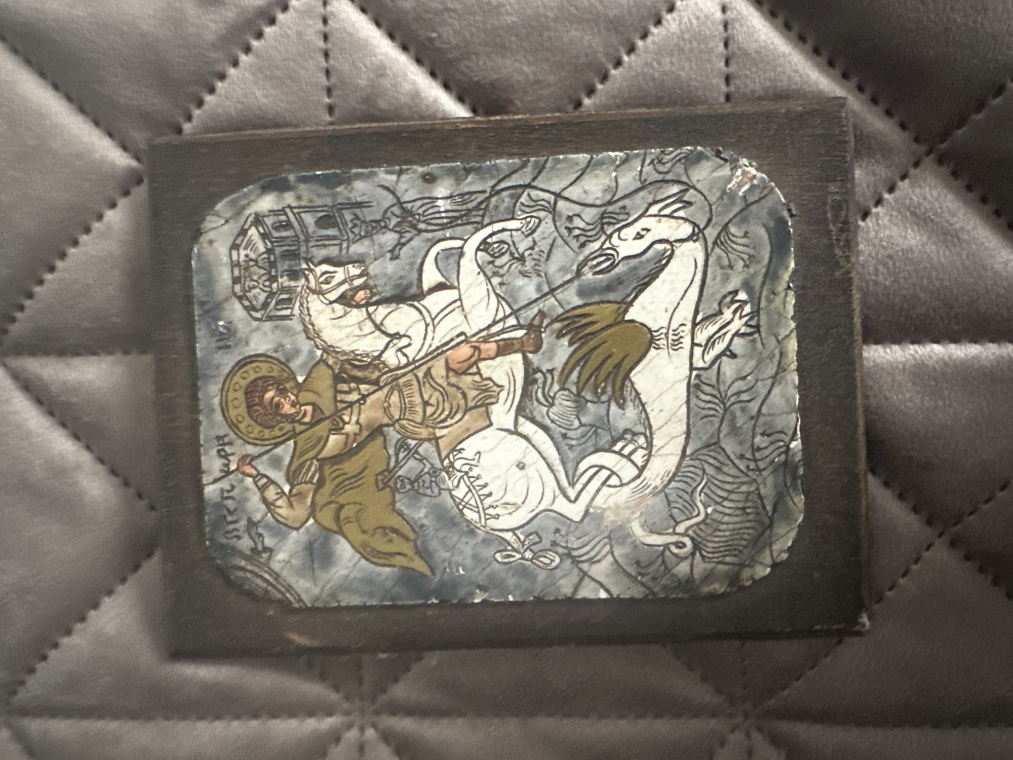 This is a close-up, top-down shot of an icon depicting Saint George slaying a dragon, mounted within a dark wooden frame. The icon is rectangular with rounded corners and appears to be made of a ceramic or enamel material, showing some signs of wear and chipping around the edges. The central figure is Saint George, dressed in a golden cloak and armor, riding a white horse. He is in the process of spearing a white dragon with dark outlining. The background is a textured gray with some abstract, dark wavy lines. To the right of Saint George, there is a multi-tiered building with figures visible in its windows. Above Saint George's head, there is text written in what appears to be Cyrillic script. The icon is placed on a dark gray, quilted surface. The image does not provide any visual information about the city of Marten, Bulgaria, or any specific location within it.