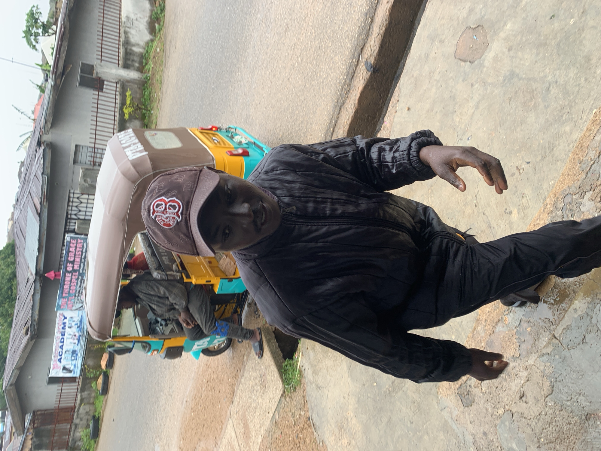 In Jos, Nigeria, a man wearing a black jacket and pants, and a brown baseball cap with a red and white logo, walks towards the camera on a wet, concrete path. Behind him, a yellow and teal auto-rickshaw is parked on the side of a street. The auto-rickshaw has a brown canopy and a banner on its side that reads "THE THRONE OF GRACE GOSPEL MINISTRY". Another banner above the auto-rickshaw reads "ACADEMY". The scene appears to be in a residential or commercial area with buildings and fences visible in the background. The ground is wet, suggesting it has recently rained.
