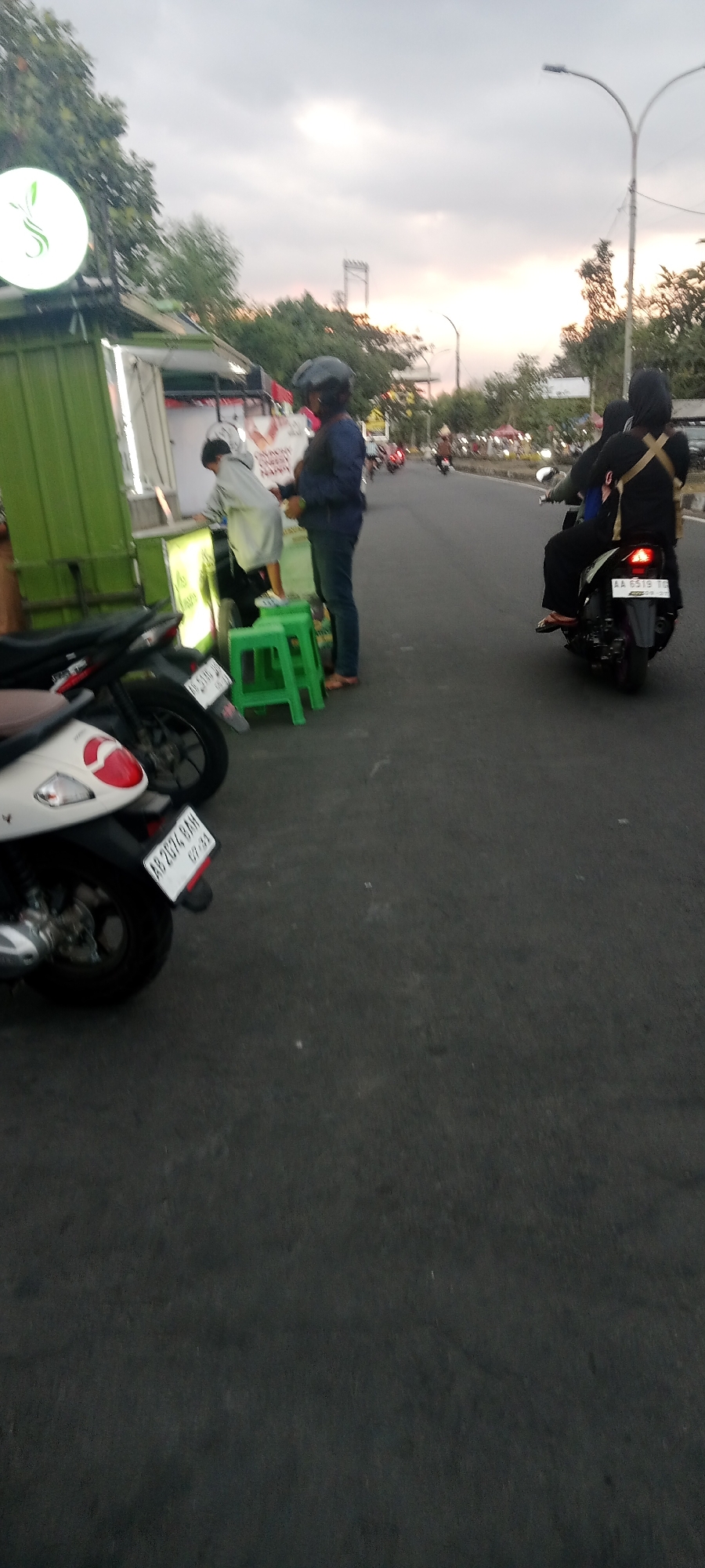 A full shot captures a street scene in Ngemplak, Indonesia, under an overcast sky at dusk. On the left, a green food stall is illuminated by internal lighting, with a white circular sign featuring a green stylized leaf logo. A person in a light grey jacket, possibly a customer, stands at the counter. Next to them, a person in a dark blue shirt and jeans, wearing a black helmet, stands with their back to the viewer. Two green plastic stools are placed near the food stall. In the foreground, the rear of two scooters is visible; one is white with a red tail light, and the other is darker with a white license plate reading "AB 2074 BAN". Further down the asphalt road, a black motorcycle carries two individuals wearing dark clothing and headscarves, with the rear passenger visible. Several other vehicles and people are present on the road in the distance, along with more stalls or shops lining the street. A streetlamp with a curved arm is visible on the right side of the frame. The overall scene suggests a bustling evening atmosphere in a semi-urban area.