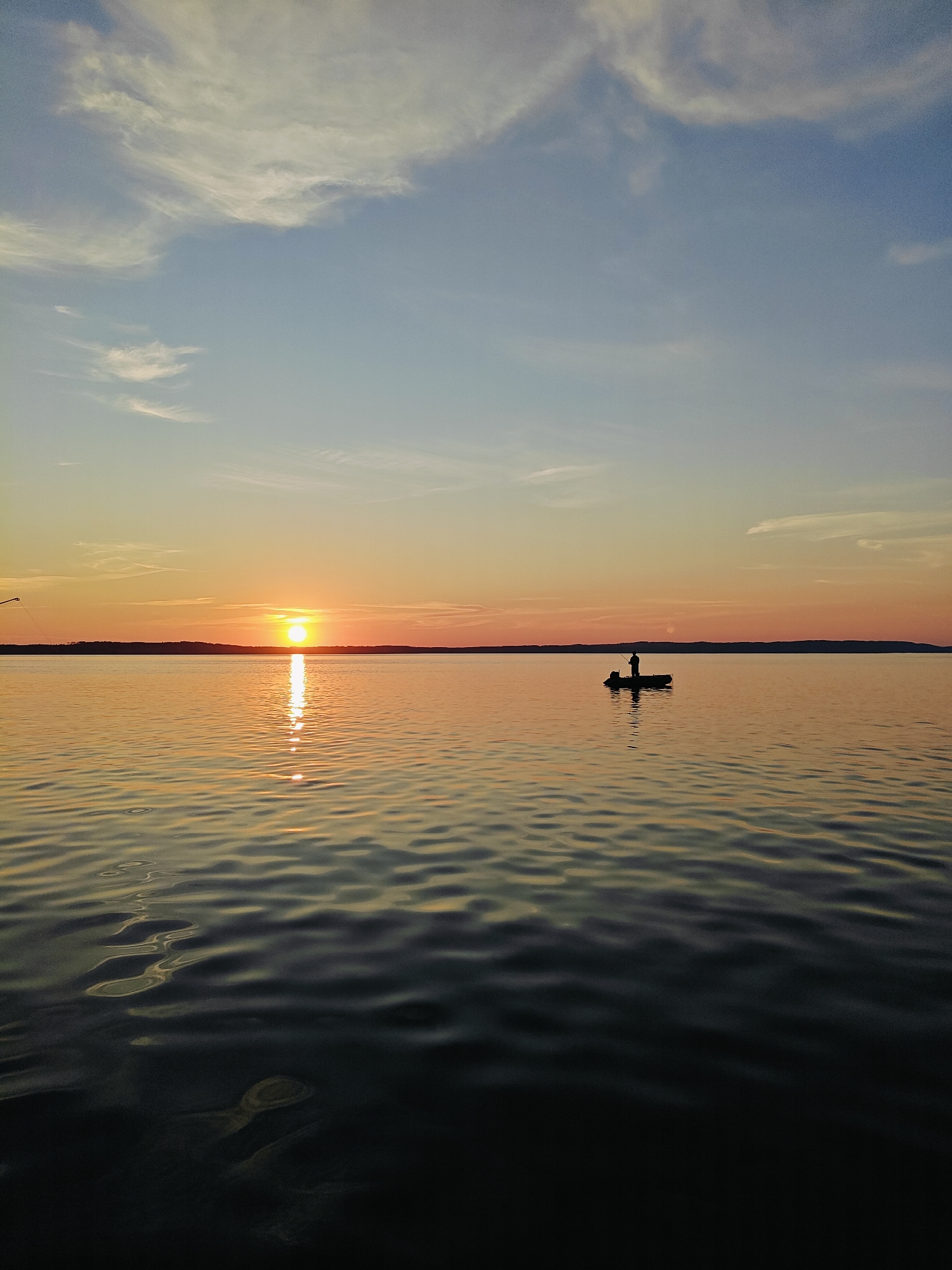 A lone fisherman stands in silhouette on a small boat in the middle of a calm body of water. The sun is setting on the horizon, casting a warm orange glow across the sky and reflecting on the water's surface. The sky is a gradient of blue and orange, with scattered wispy clouds. The water is dark and rippled, with the sun's reflection creating a shimmering path. The scene appears to be a serene and peaceful evening in Chaykovsky, Russia.