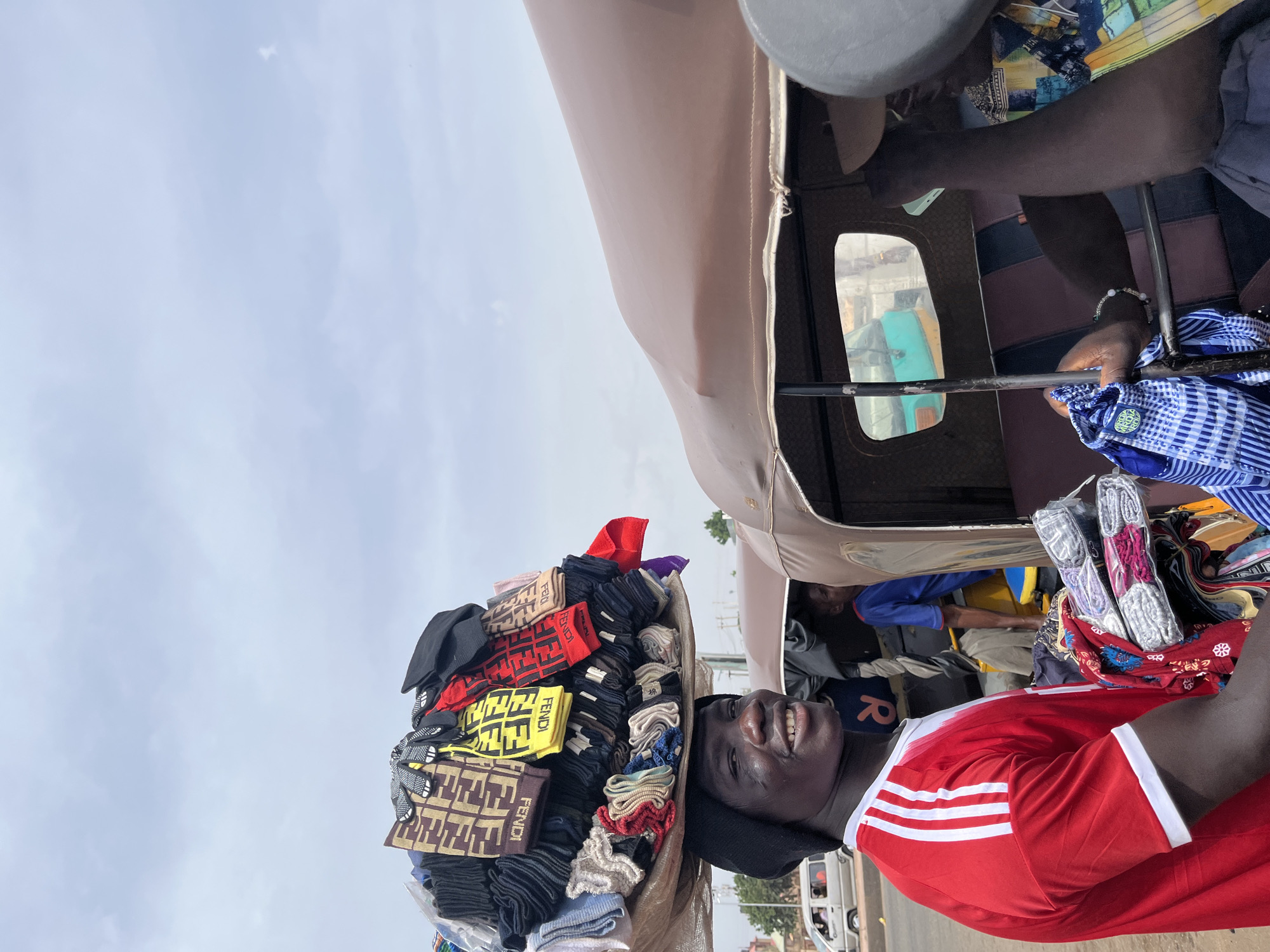 A man in a red shirt with three white stripes on the sleeve is standing in front of a vehicle with a beige roof. On his head is a large stack of Fendi-branded socks and hats. Another individual can be seen inside the vehicle. The scene appears to be taking place on a street in Gura Top, Nigeria, as evidenced by the presence of distinctive local transportation and architectural styles.