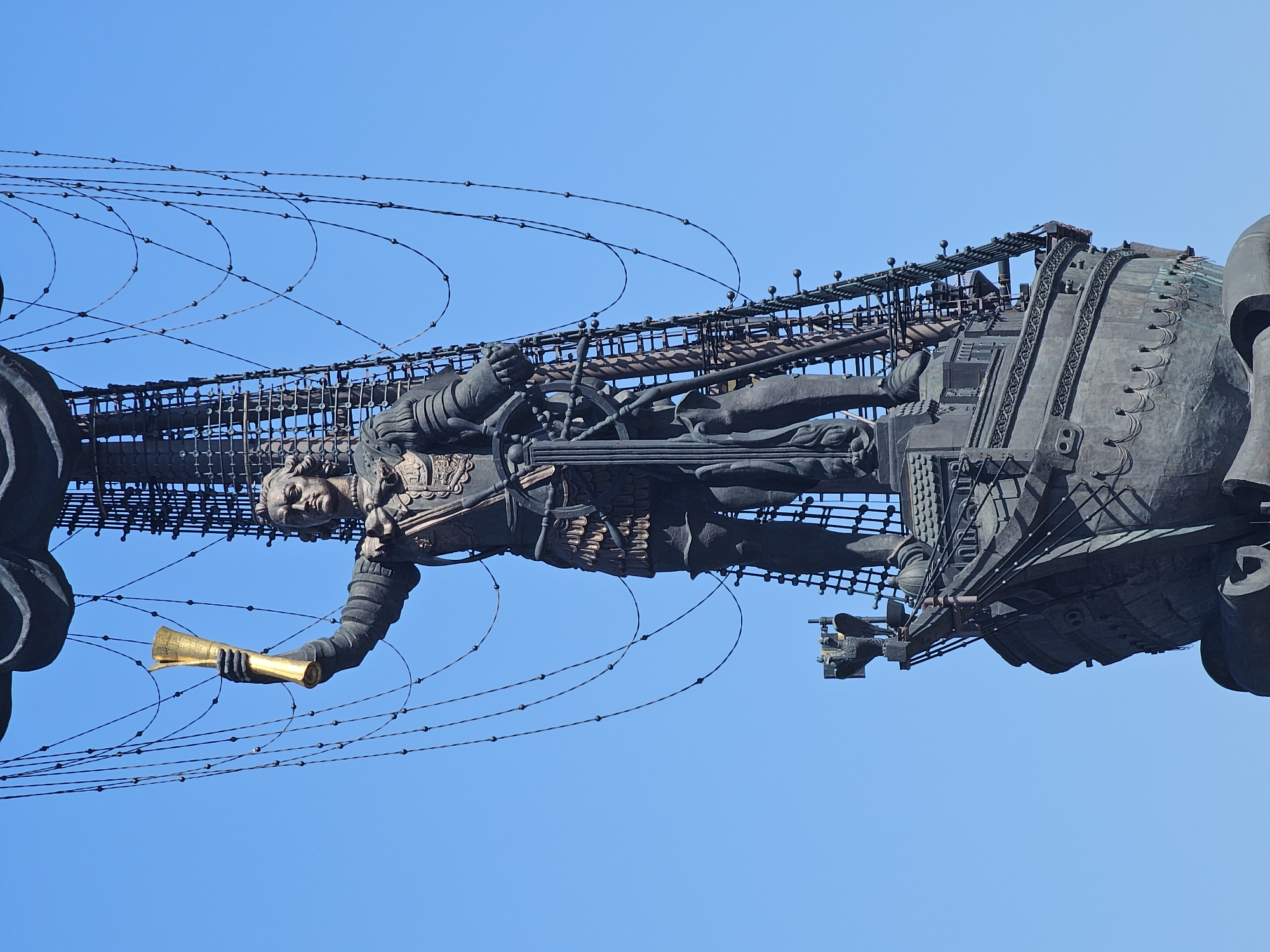 The image depicts a statue of Peter the Great in Moscow, Russia, set against a clear blue sky. The statue is made of bronze and features Peter the Great standing in a stylized ship-like structure. He is holding a golden scroll in his outstretched hand. The statue is supported by a metal framework of wires and rods. The overall composition suggests a dynamic, towering presence.