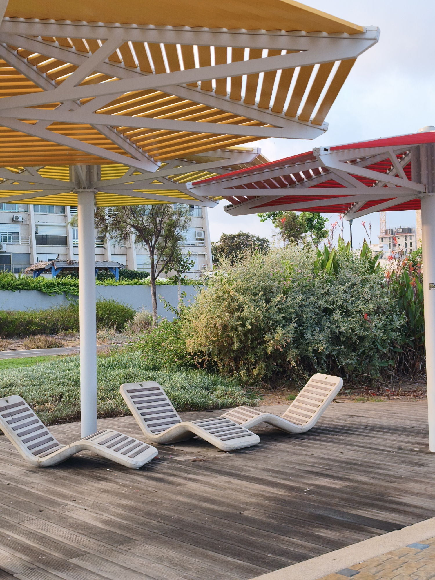 The image shows a public area with several modern lounge chairs on a wooden deck. Above the chairs are two large, colorful sunshades, one yellow and white, and the other red and white. Behind the lounge chairs is a lush green garden with various plants and trees. In the background, there is a building with balconies and windows, typical of residential architecture in coastal cities. There are also some construction cranes visible in the distance, suggesting ongoing development in the area. The sky is overcast, indicating it might be early morning or late afternoon. The scene appears to be a relaxing outdoor space, possibly a park or promenade in a city like Nahariyya, Israel, known for its Mediterranean coastline and pleasant climate. There are no people visible in the image.