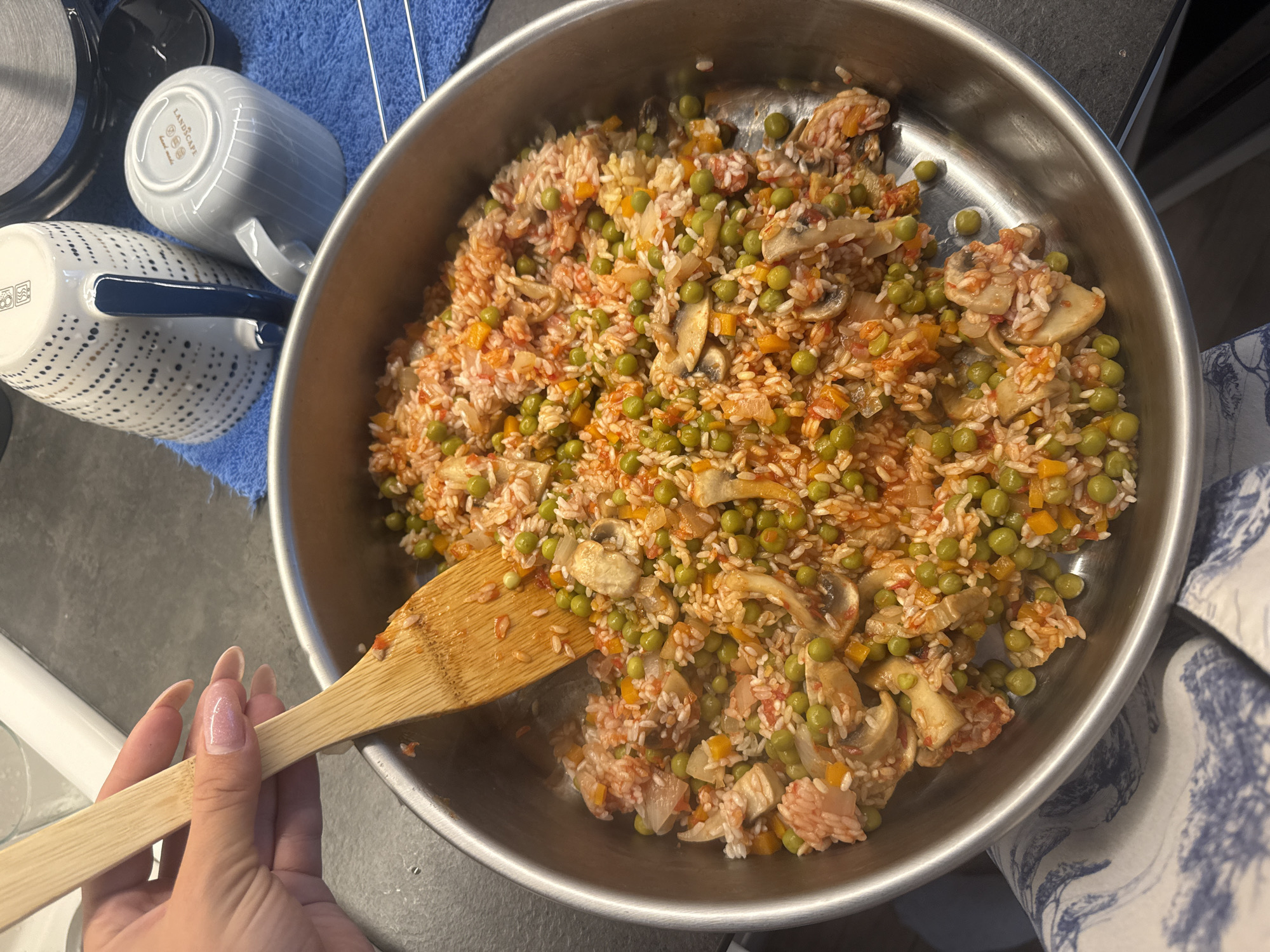 An overhead shot captures a home kitchen scene focused on a pan of cooking food. A large, round, silver metal pan occupies the central and right portions of the image, filled with a colorful rice dish. The meal consists of white rice mixed with bright green peas, sliced light brown mushrooms, and diced orange and red vegetables (likely carrots, bell peppers, and tomatoes), giving it a vibrant appearance.
A wooden stirring spoon is partially submerged in the food, and a hand, presumably female, with long, light pink manicured nails, holds the spoon's handle from the lower-left side, suggesting active cooking or mixing.
To the left of the pan, on a dark gray countertop, two ceramic mugs are visible. They rest on a blue cloth, possibly a dish towel. The closer mug is white with blue dots and a blue handle. Its base shows care symbols for being dishwasher and microwave safe, along with the number "22212." Behind it, a second white mug with light gray stripes also displays text on its bottom, including the word "LANDSCAPE" and other smaller, unreadable details. A white and blue patterned textile, possibly a kitchen towel or oven mitt, is partially visible on the right side of the pan. The scene depicts the preparation of a meal in a domestic kitchen setting. No specific time of day or location is discernible.