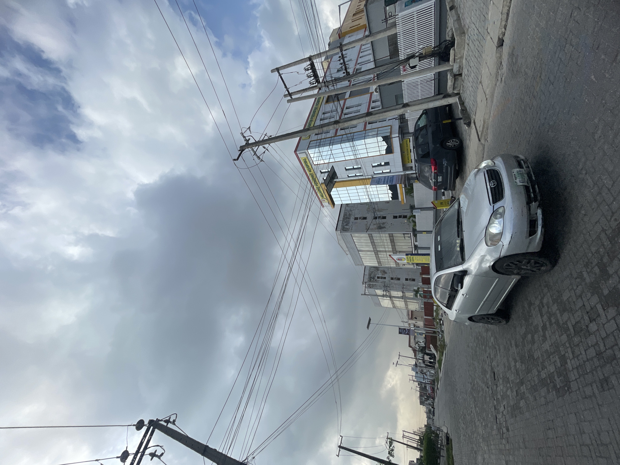 The image shows a street scene in Moba, Nigeria. The focus is on a silver car parked on a brick-paved road. A dark-colored minivan is parked behind the silver car. Several buildings are visible along the street, including a multi-story building with reflective windows and what appears to be commercial signage. Utility poles and overhead power lines are prominent in the scene, crisscrossing the sky. The sky is overcast with varying cloud cover. Other cars are visible in the distance, indicating moderate traffic. No people are clearly visible. The environment is urban.