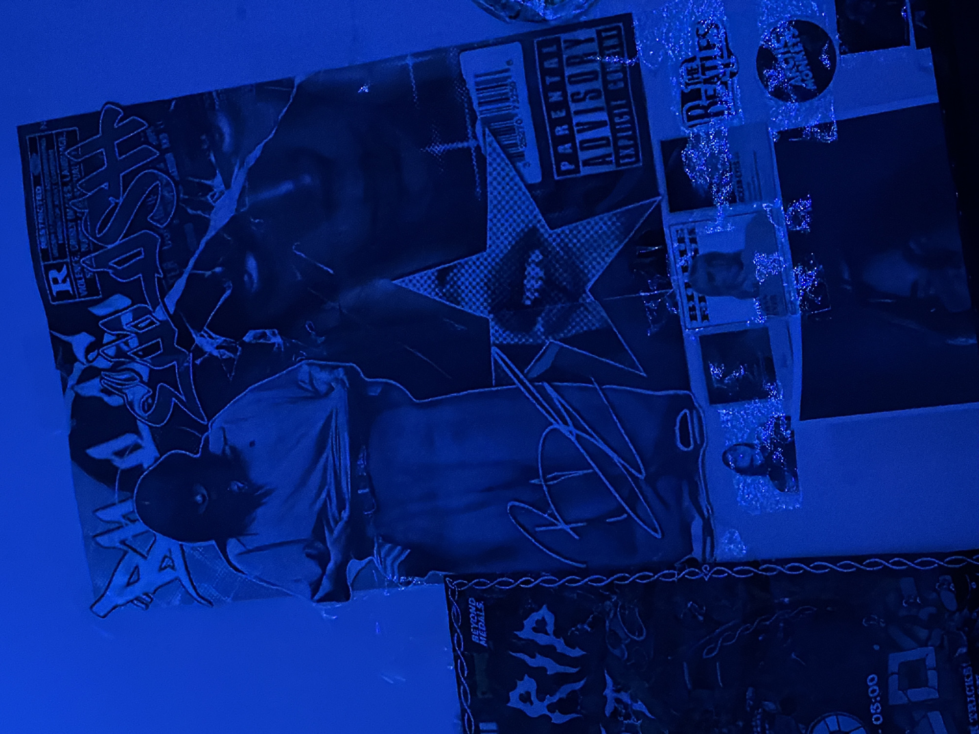 The image depicts a dimly lit wall adorned with various posters and photographs, all bathed in a strong blue light. The primary focus is a large poster featuring a figure in a black balaclava and a light-colored shirt, holding it up. Above this figure, text reads "BAD LUSH" in a stylized font, and to the right, a "PARENTAL ADVISORY EXPLICIT CONTENT" label is visible.
To the left of the main poster is another poster with the text "RIP" and "BEYOND MEDALS." Below the main poster are several smaller images, including what appear to be album covers or photos of musicians. Visible text on these smaller images includes "THE BEATLES" and "ARCTIC MONKEYS."
The overall impression is that of a personal collection of music or art displayed on a wall. The blue lighting creates a dramatic and somewhat abstract atmosphere. There is no discernible context for this scene to be located in Oslo, Norway.