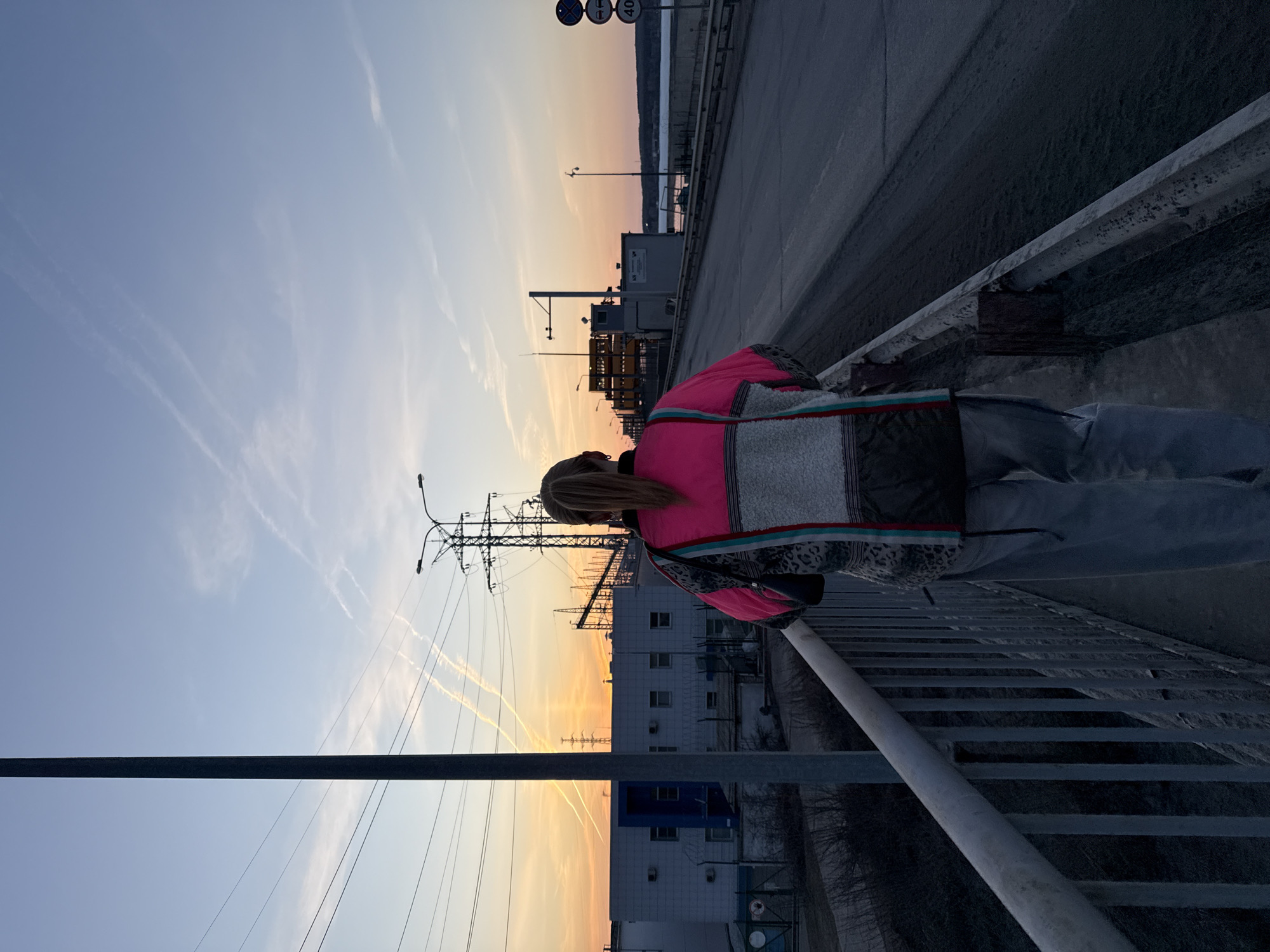 A person is leaning over the railing of a bridge overlooking the city of Chaikovsky, Russia, during a sunset. The person is wearing a pink, grey, and black jacket with light grey pants. The bridge has grey concrete railings. In the background are buildings, a body of water, power lines, and traffic signs. The sky is multi-toned with streaks of pink, orange, and blue. Contrails are visible in the sky.
