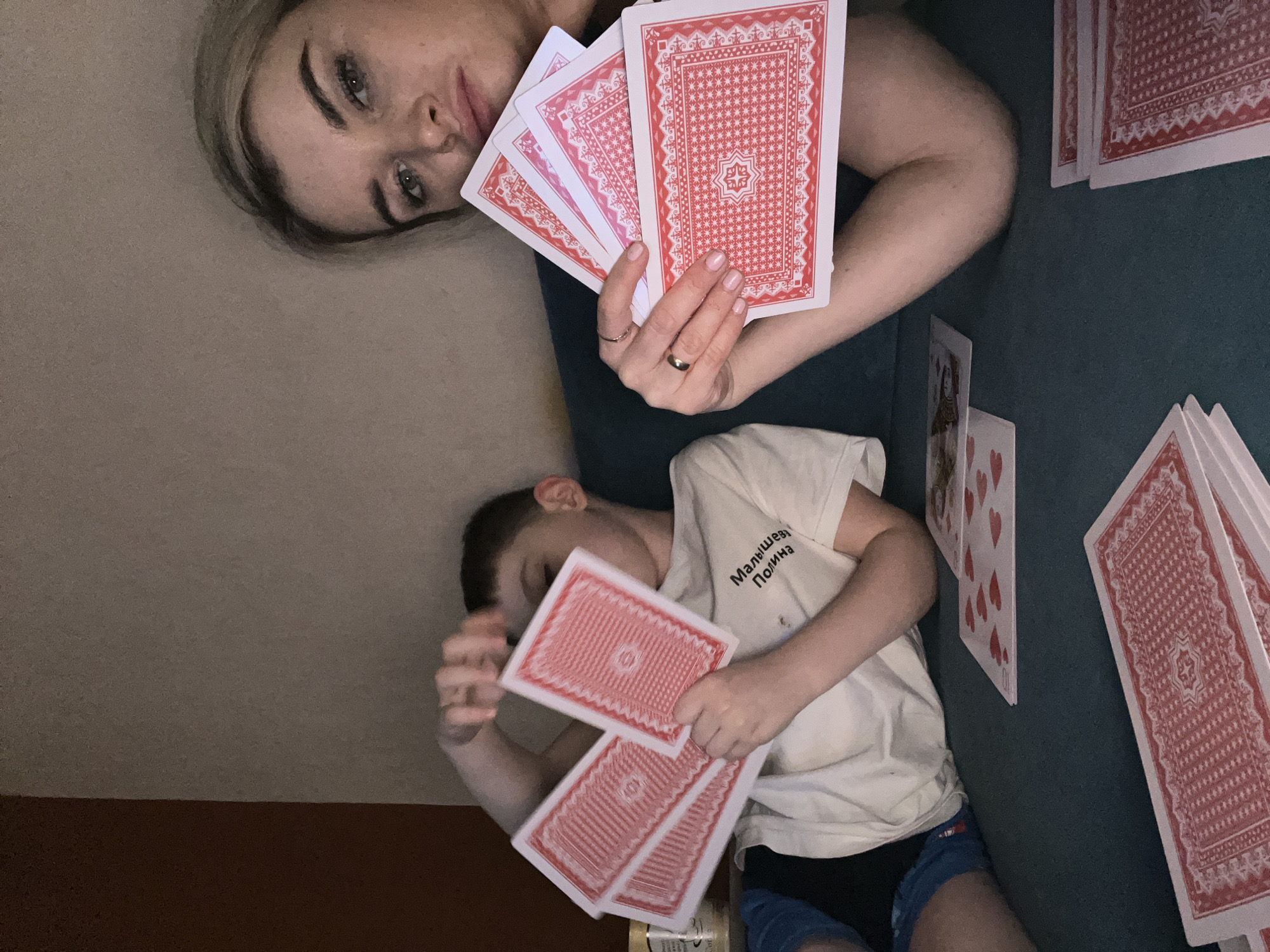 The image captures a relaxed indoor scene featuring a woman and a young boy engaged in playing cards. The woman is positioned on the left, looking directly at the camera with a calm expression. She has light-colored hair and is wearing rings on her fingers, one of which appears to be a wedding band. She holds a fan of red-backed playing cards in her left hand, displaying the patterned reverse sides.
To the right, a young boy lies on a dark green or teal surface, likely a couch or bed, holding several red-backed playing cards in his hands. His face is partially obscured by his arm and the cards. He is wearing a white t-shirt with black text in Cyrillic, which reads "Малышева Полина" (Malysheva Polina), and blue shorts.
Around the individuals, more playing cards are scattered on the green surface, some face-down showing their red backs, and others face-up revealing hearts and what appears to be a queen card. The background consists of a plain light brown or beige wall and a darker brown lower section, possibly a floor or baseboard. The lighting suggests an indoor setting, possibly in the evening or with artificial light. The overall atmosphere is one of leisure and family activity in Chaikovsky, Russia.