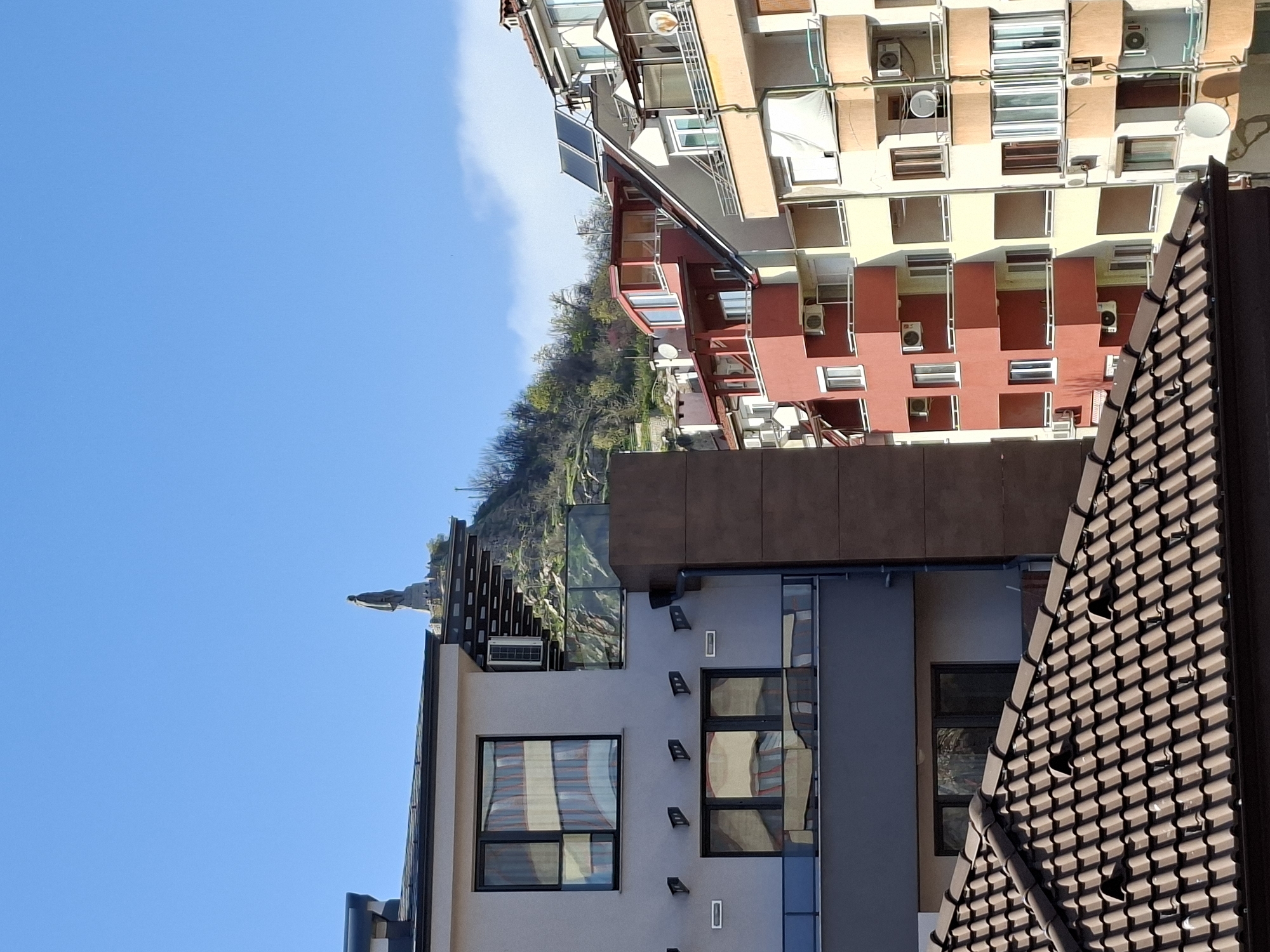 The image shows an upward view of buildings in Plovdiv, Bulgaria, against a clear blue sky. A modern building with a light gray facade and dark-framed windows is in the foreground. A brown tile roof is partially visible on the right. A statue is silhouetted against the sky atop the building. Behind the modern building is a tree-covered hill. In the background, there is a taller, multi-story building with a mix of beige and red painted sections. The building has many balconies and air conditioning units are visible.