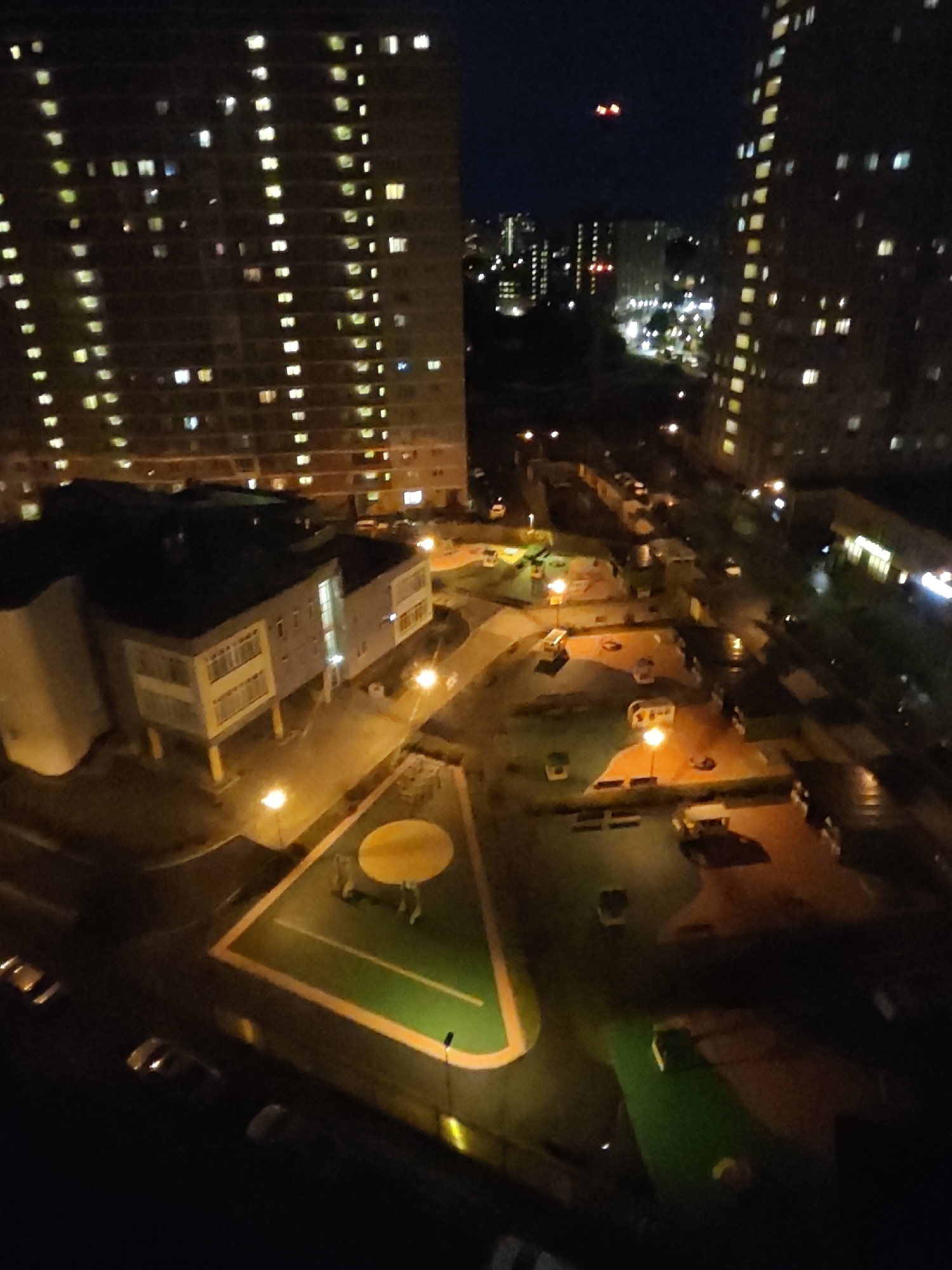 At night, tall, illuminated apartment buildings are visible in the background, with lights on in many of the windows. In the foreground, there is a playground area with a yellow circular structure and some equipment. A low-rise building, possibly a kindergarten or community center, is situated between the apartment buildings and the playground. There are also streetlights illuminating the area and several cars parked along a street. The sky is dark blue, indicating it is nighttime. The scene is set in the city of Shcherbinka, Russia.