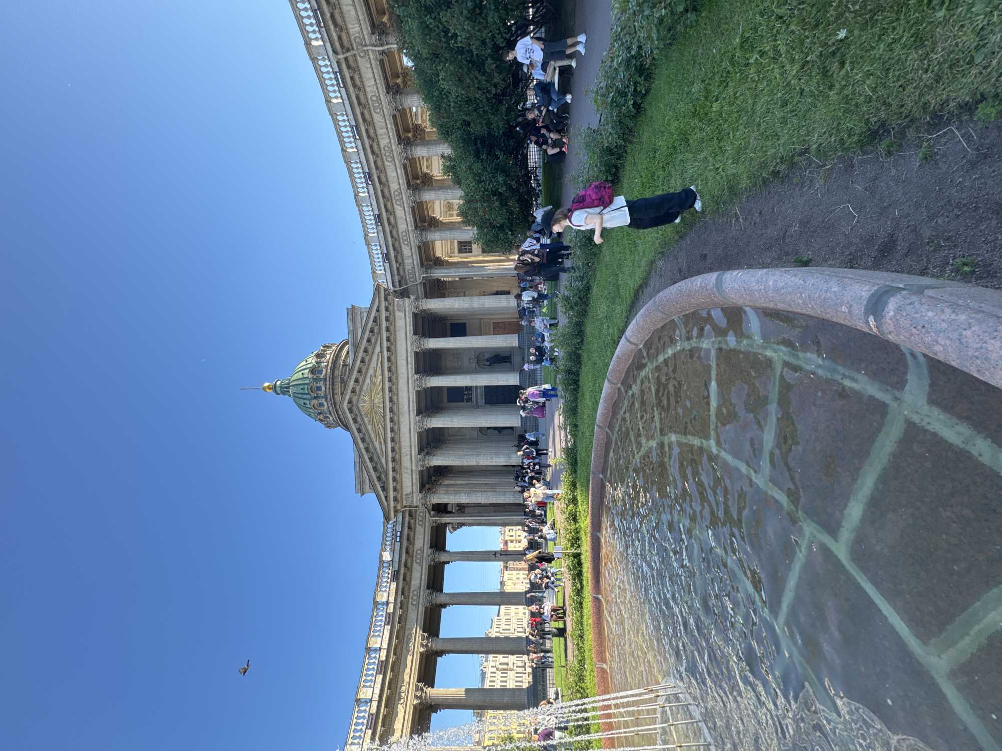 The image shows a wide, outdoor, sunny view of Kazan Cathedral in Saint Petersburg, Russia. In the foreground, a large, circular fountain with clear, rippling water is visible on the left. The fountain's edge is made of pinkish stone. A person wearing a white t-shirt, black pants, and a pink backpack is standing to the right of the fountain, facing away from the camera and looking towards the cathedral. Beyond the fountain and the person, a green lawn and bushes are present. In the background, the grand Kazan Cathedral stands with its distinctive curved colonnade and a green dome topped with a golden spire. Numerous people are scattered across the plaza in front of the cathedral, some sitting on benches, others walking, and some gathered in groups. The sky is a clear, bright blue. A single bird is captured in flight in the upper left quadrant of the image.