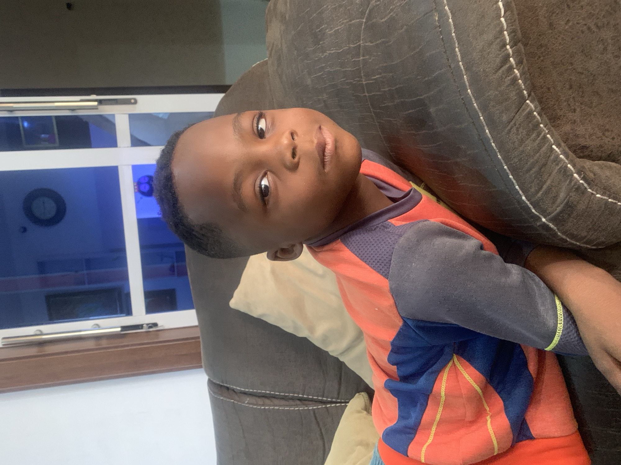 A young, dark-skinned boy is lying on a brown couch with a light-colored pillow. He's wearing a multi-colored shirt with gray sleeves, red torso, and blue patches. The boy is looking at the camera with a slight smile. Behind him is a window with blue-tinted glass, likely reflecting the sky. The room's walls are white, and part of a clock or other circular object is visible on the wall beyond the window. There's no definitive evidence to confirm or deny that the location is Jos, Nigeria. The image does not explicitly provide information to confirm a specific city or country.