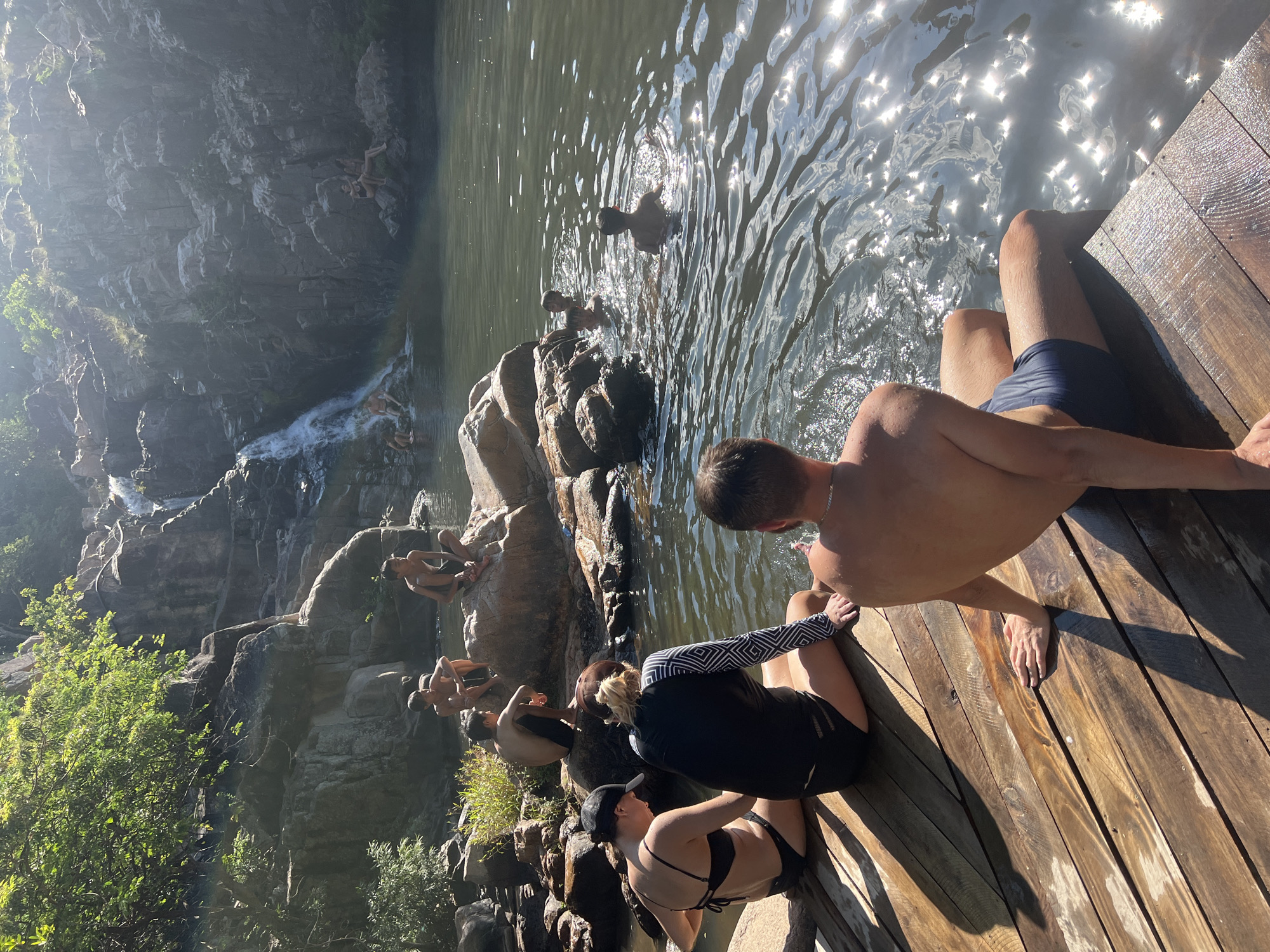 The image depicts a group of people at a natural waterfall and pool area, likely in Vĩnh Hải, Vietnam. In the foreground, two individuals are seated on a wooden platform, facing away from the camera and towards the water. To the left, a person in a black bikini and a black baseball cap is seated. Next to them, a person wearing a black long-sleeved rash guard with a geometric pattern and black bottoms is also seated. Further to the right, another individual with their back to the camera is seated on the wooden platform, wearing navy blue swim trunks.
In the water, several people are visible. Some are swimming or wading in the pool, while others are sitting on rocks near the edge of the pool or at the base of the waterfall. The waterfall cascades down a rocky cliff face, surrounded by lush green vegetation at the top. The water is relatively still, with sunlight reflecting off its surface, creating a shimmering effect. The environment appears to be a natural, outdoor setting with rock formations, trees, and a body of water. The overall atmosphere suggests a leisure activity, possibly swimming or relaxing in a natural landscape.