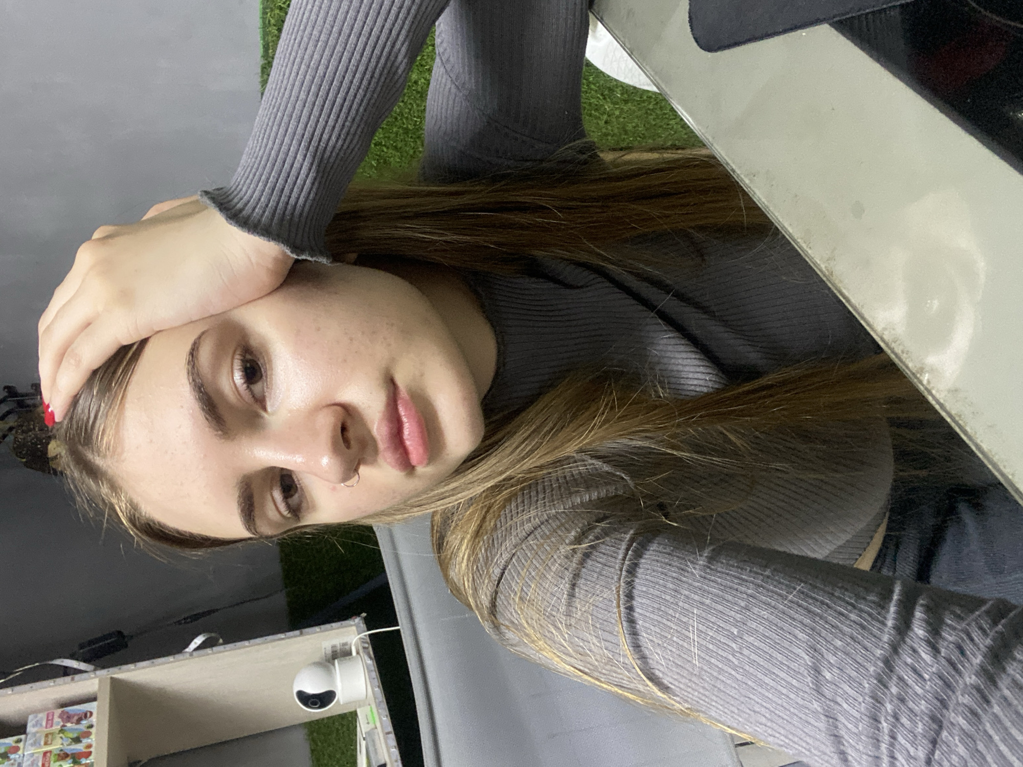 A young woman with long, light brown hair, wearing a grey ribbed turtleneck sweater, rests her forehead with her right hand. She has a silver nose ring and appears to be indoors, possibly in an urban setting. In the background, a white shelf unit is visible with various items, including what appears to be a small security camera and a grassy artificial turf surface. The overall environment suggests a casual, indoor space. The location is not identifiable as Дьяково, Russia, based on the visual information provided.