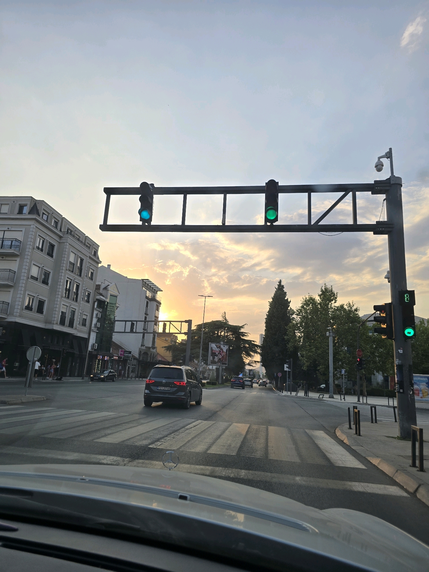 This image captures a street scene in Podgorica, Montenegro, during a sunset. A car is stopped at a crosswalk in the foreground, and a dark-colored SUV is moving away from the camera further down the street. Traffic lights are visible, with the one on the left illuminated green. Buildings line the street, some appearing to be commercial establishments with signage in a language that may be Montenegrin. Trees are present, and a security camera is mounted on a pole with the traffic lights. The sky shows a warm glow from the setting sun, with some clouds visible. The overall impression is of an urban environment at the end of the day.