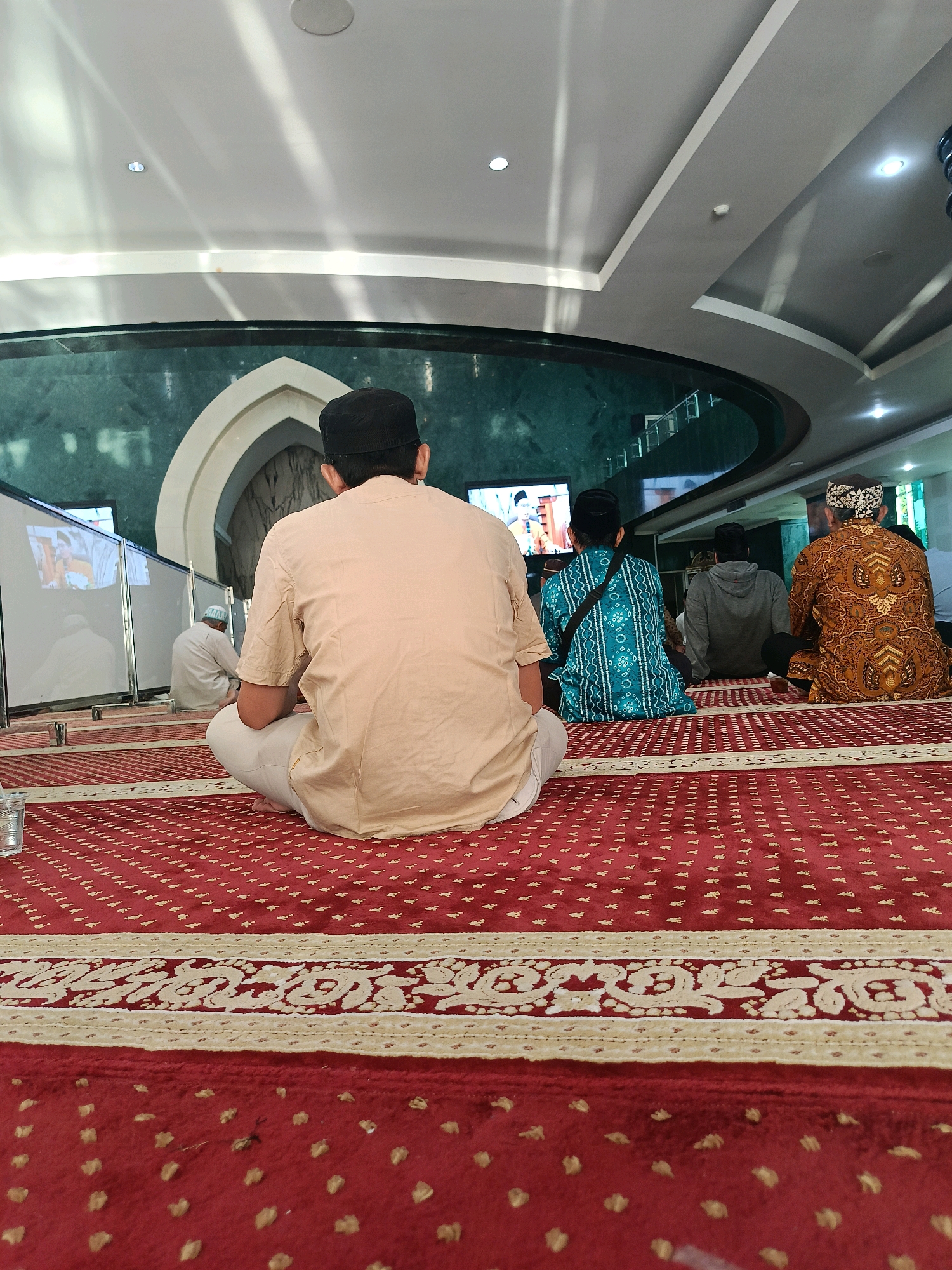 In Banguntapan, Indonesia, at the Grand Mosque of Mataram, several individuals are seen sitting on a red carpet within the mosque. The carpet features intricate patterns in beige and red. The individuals are facing away from the camera, with the closest person wearing a light brown, collared shirt and white pants, and a black peci (cap). To the left, another person in white is also seated, with a screen displaying an image visible behind them. Further to the right, two more individuals are seated: one in a blue patterned shirt with a black strap across their chest, and another in a brown patterned garment. The background reveals a curved, dark green wall and an arched architectural feature. Above, the ceiling is a combination of white and grey panels with lighting fixtures. The overall scene depicts a moment of prayer or contemplation within a mosque setting.