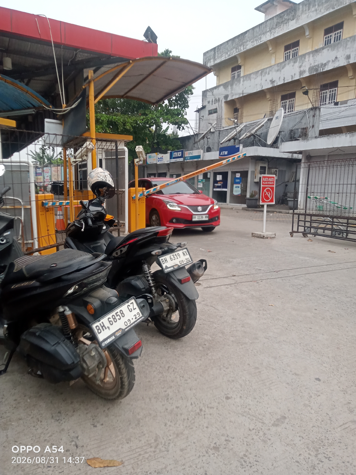The image depicts an outdoor scene at a gated entrance, likely a commercial or residential area, with elements suggesting it is located in Jambi, Indonesia. Two motorcycles are parked in the foreground, one black and one dark colored, with their license plates visible: "BH 6858 CZ" and "BH 6319 NX". A red Suzuki car is seen approaching the gate, which is partially lowered. The gate area features yellow and white striped barriers, signage including "ATM" and "HATI-HATI DILARANG OTOMATIS," and various security cameras and satellite dishes mounted on the adjacent building. The building itself is a light-colored, multi-story structure with numerous windows. The ground is paved concrete. The overall atmosphere suggests a typical urban scene in Indonesia.