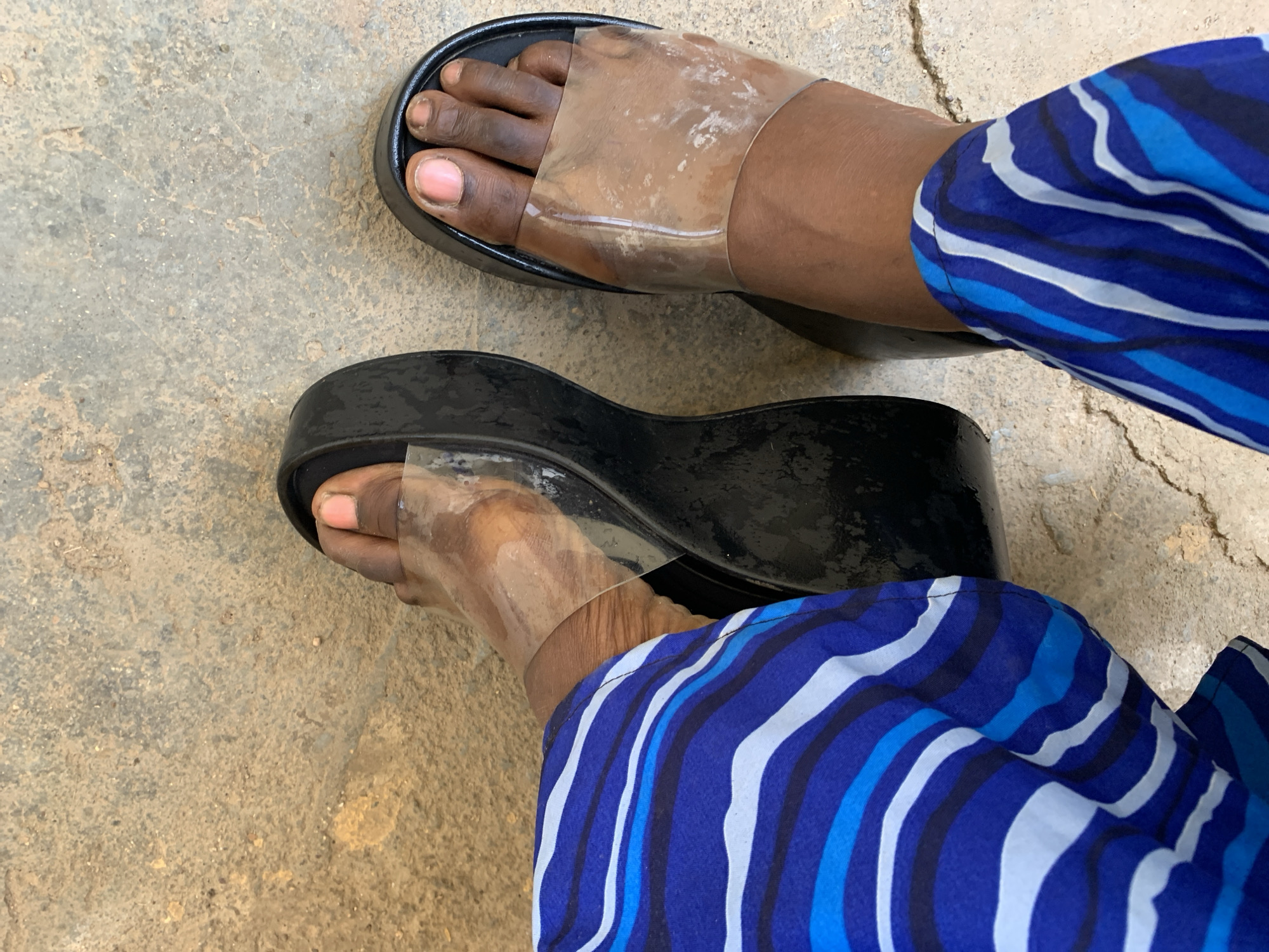 A top-down medium shot depicts a person wearing platform sandals with clear straps, standing on a concrete surface. The person's feet, toes visible under the clear straps, are adorned with the black platform sandals. A blue and white striped garment covers part of their lower leg. The concrete surface appears slightly uneven and textured. The location is not discernible from the image. There is no specific information indicating the city of Rafin Par, Nigeria.