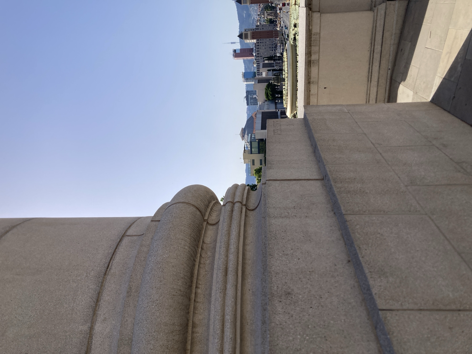 The image captures a panoramic view of Barcelona, Spain, taken from a high vantage point. In the foreground, a large, textured stone column, likely part of a monumental structure, dominates the left side of the frame. The column exhibits classical architectural details with multiple carved bands. To the right, a wide expanse of blue sky stretches across the upper portion of the image.
Below the sky, the cityscape of Barcelona unfolds. A circular fountain with illuminated jets is prominently featured in the mid-ground, surrounded by a well-manicured area. Beyond the fountain, a dense urban landscape of buildings rises, showcasing a variety of architectural styles and heights. Notable structures include modern high-rises with glass facades and older buildings with distinct architectural features, such as the twin towers that flank a distant street. A prominent telecommunications tower is visible on the distant hills.
The lower right section of the image reveals a paved plaza with large stone tiles, casting distinct shadows from an unseen light source, suggesting it is daytime. The overall scene is bathed in bright sunlight, indicative of a clear day. No people or animals are visible in the image. The composition offers a juxtaposition of grand, classical architecture in the foreground against the vibrant, modern urban fabric of Barcelona.