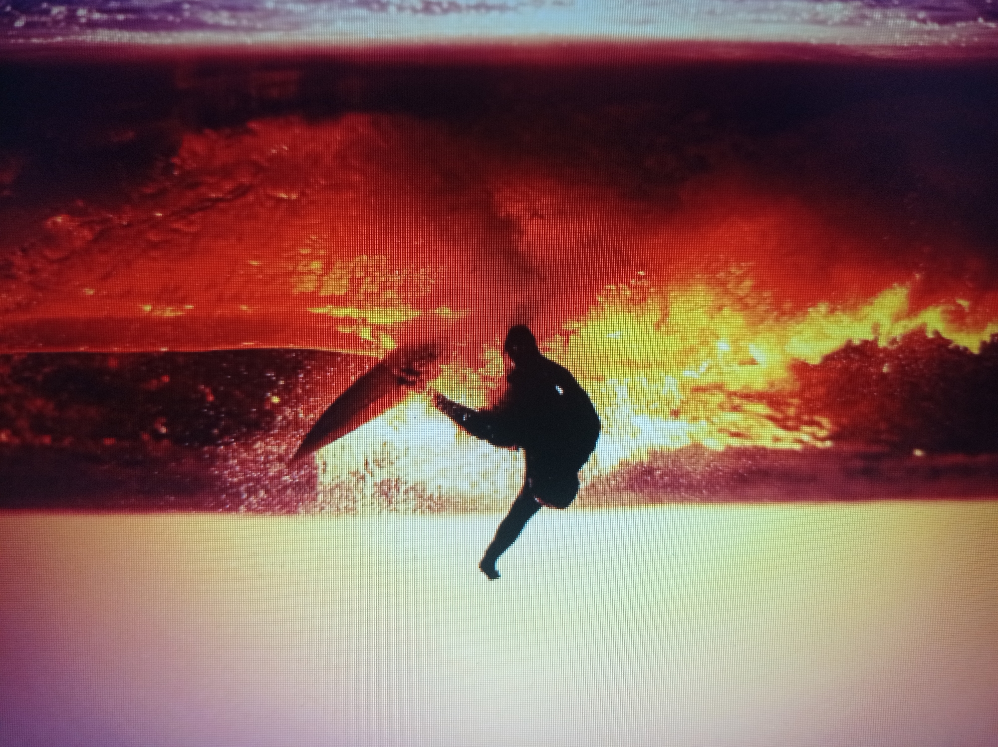 The image is a low-resolution, pixelated photograph of a surfer silhouetted against a bright sky and ocean waves. The surfer is in mid-air, appearing to perform a maneuver on a surfboard. The water is depicted with vibrant oranges and yellows, suggesting either a sunset or sunrise. The sky transitions from a pale yellow at the horizon to a lighter hue towards the top. The overall scene is dynamic and captures a moment of action in the ocean. There is no information in the image to suggest a location in the city of Mukachevo, Ukraine.