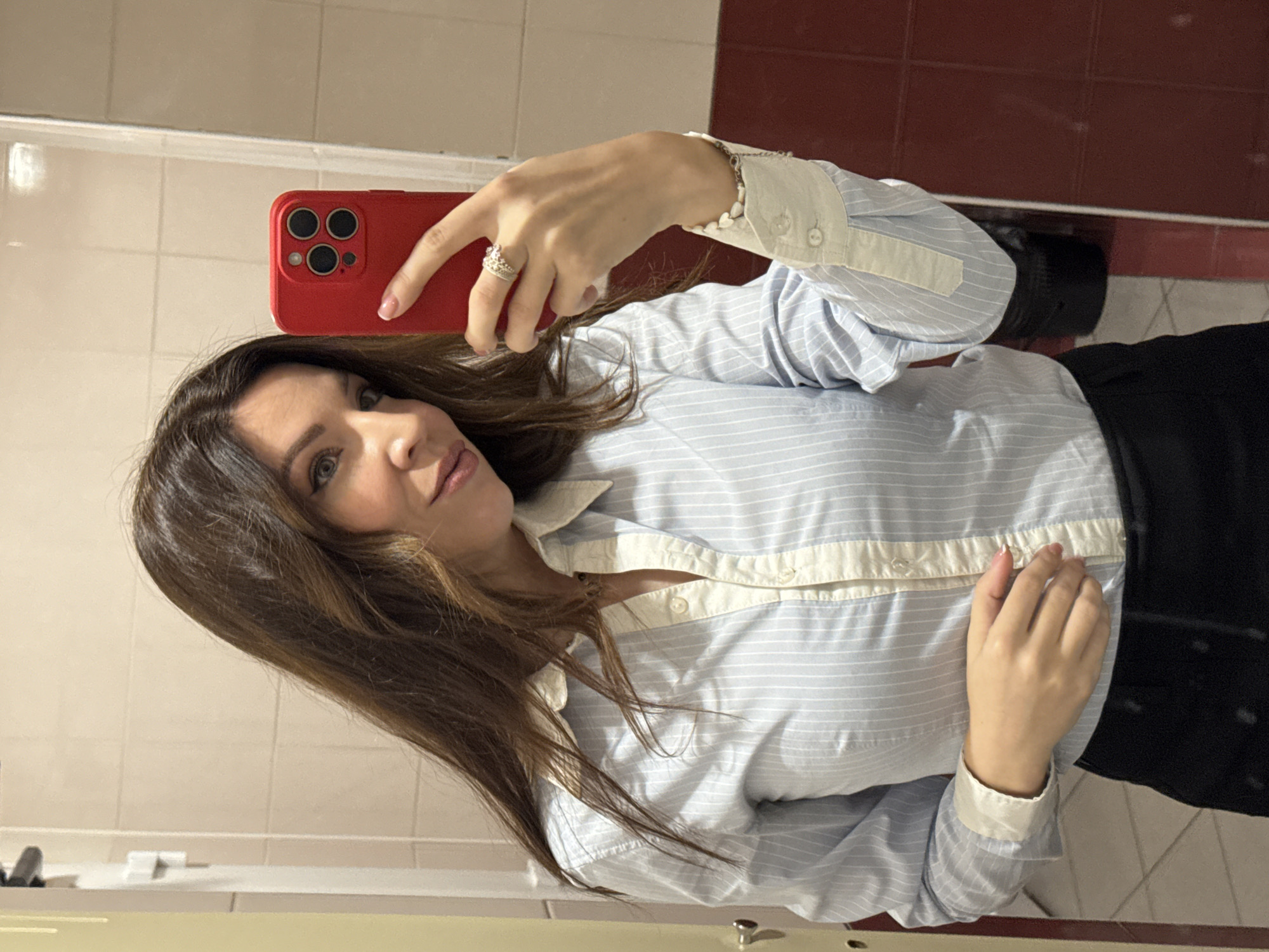 A woman is taking a selfie in a bathroom. She is holding a red phone with her left hand and looking at the screen. She is wearing a light blue and white striped button-down shirt, a ring on her ring finger, and a bracelet. Part of a black garment is seen on the right side of the image. The background consists of beige and white tiled walls, a mirror reflecting the camera, and a red tiled wall on the upper right. The location is Unknown, Unknown.
