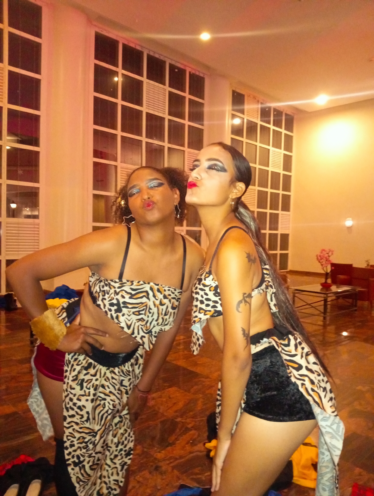 Two young women stand indoors. They are dressed in matching outfits with leopard-print tops and bottoms, with a black belt. They are each pursing their lips in a kissing gesture, as if posing for a photograph. The woman on the left has her hand on her hip. The woman on the right has her hands resting on her thighs. Both have elaborate eye makeup. A window with a grid pattern is visible in the background. There are also chairs and a table with flowers in the background. The location is likely Pedro González, Venezuela.