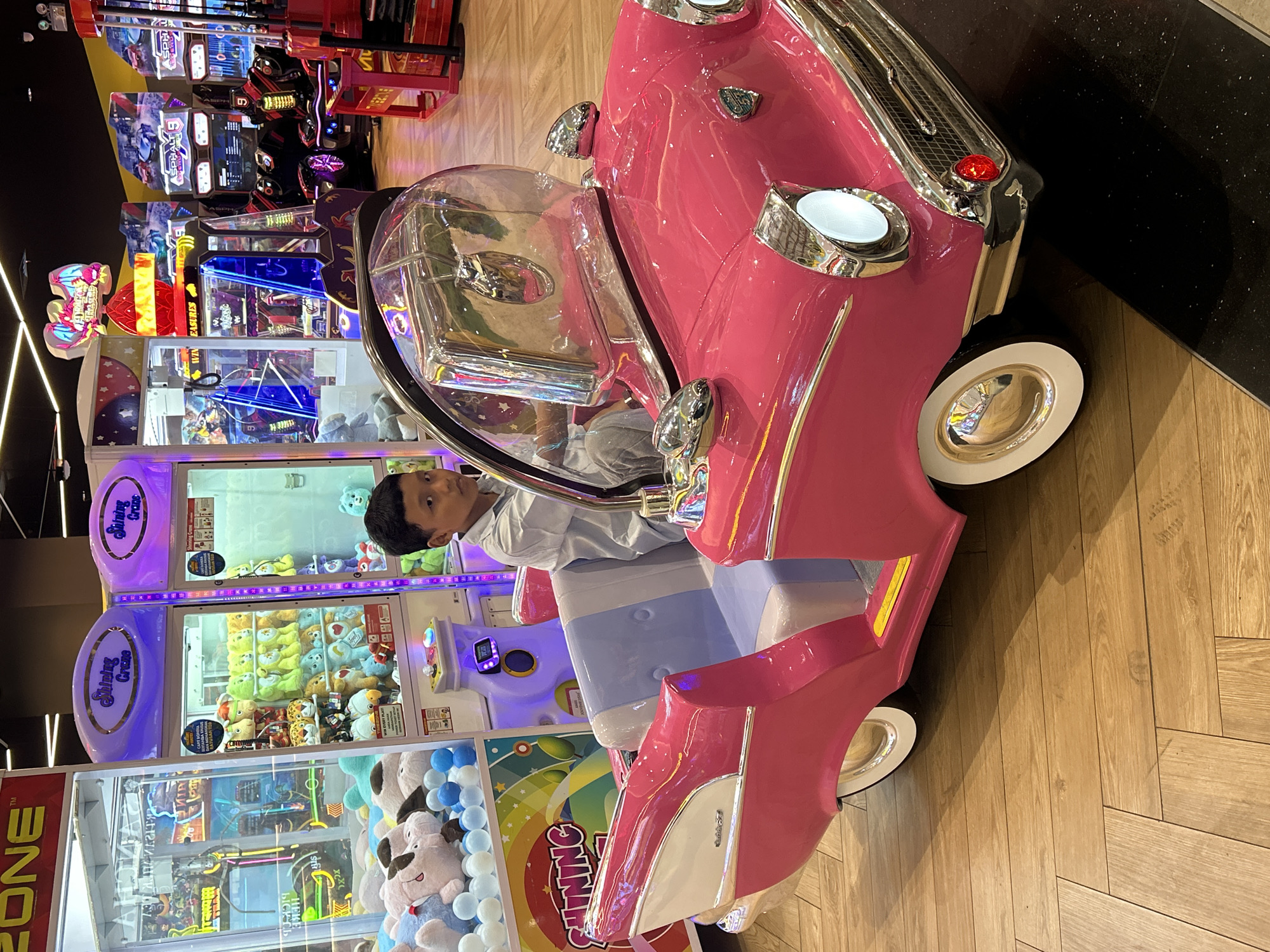 A young boy sits in a pink, vintage-style ride-on car at an arcade in Makassar, Indonesia. He is wearing a light blue collared shirt and grey pants. The car has a clear windshield and chrome accents. In the background, several arcade machines are visible, including claw machines filled with plush toys and racing games. The floor is made of light brown wood planks. The lighting appears to be from overhead fluorescent lights and the colorful lights of the arcade machines.