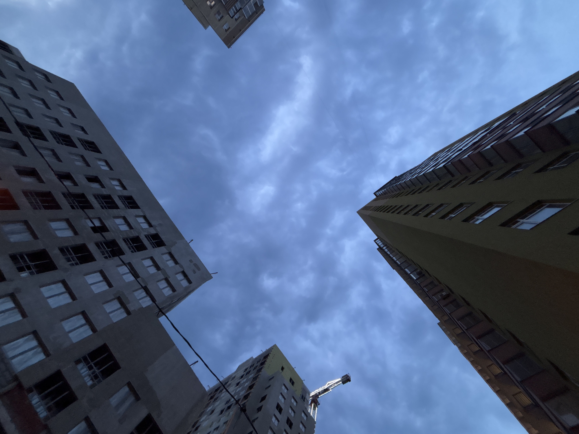 Looking up, the image captures several buildings against a cloudy sky, indicating an urban environment. The buildings are multi-story structures of varying design and construction status. One building appears to be undergoing construction, with incomplete exterior walls and a visible crane. Another building has a yellow facade with windows.
Based on the composition, the image may have been taken in the city of Perm, Russia. The city has modern, mixed-use buildings, similar to what is seen in the photo.