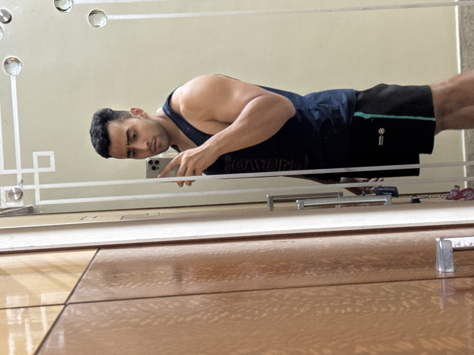 A person in a dark tank top and shorts takes a selfie in a large mirror. The mirror has a decorative design with circles and linear patterns. The person holds a phone and looks directly at it. The bottom of the mirror is resting on a wooden surface with tiled segments. The reflection shows the person's muscular build. The angle of the shot suggests the person is lying on the floor taking the picture.
