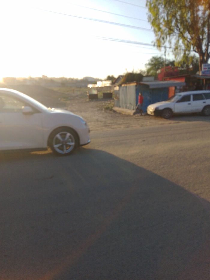 The image shows a street scene in Athi River, Kenya, taken from the perspective of a white car stopped on the road. The image is blurry and overexposed, likely due to glare from the sun. The car is facing towards the right of the frame. In the background, there is a small, blue building on the right. A person wearing a red shirt is walking toward the camera on the right. A white car is parked to the right of the person. The road is paved and there are some buildings visible in the distance. The scene is sunny and the light is bright. There are a few power lines running across the sky. The scene is likely taken during the late afternoon or early evening.