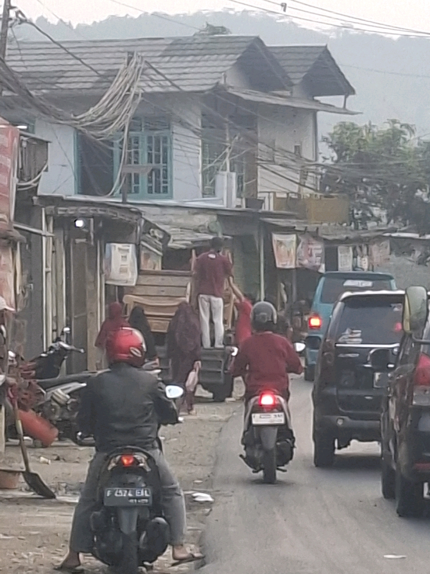 The image depicts a street scene in Cijalingan, Indonesia, with several people and vehicles. In the foreground, two individuals are riding motorcycles. The person on the left is wearing a black jacket and a red helmet, while the person on the right is wearing a red long-sleeved shirt and a dark helmet. Behind them, a blue van is visible, and further back, a dark-colored SUV is also on the road. Several buildings line the street, some appearing to be shops or houses with corrugated metal roofs and various items displayed outside. A person in a red shirt is standing on a platform behind a motorcycle, and another person in a dark garment is standing beside them. The overall environment suggests a residential or commercial area with moderate activity. The sky is overcast, and the lighting is diffused, indicating either early morning or late afternoon. There are also utility wires visible above the buildings.