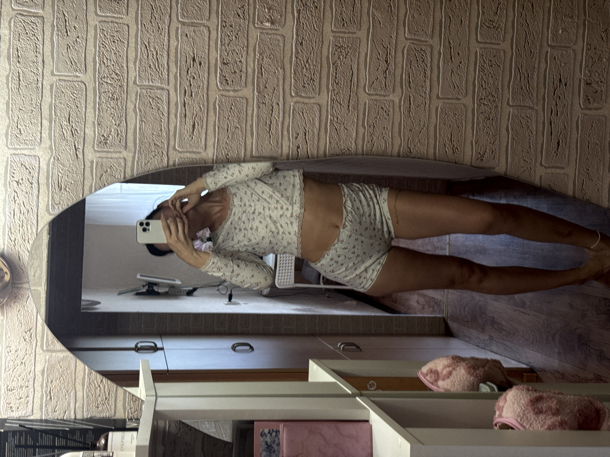 A woman is taking a selfie in a mirror. She is wearing a light-colored pajama set with a floral pattern, consisting of a long-sleeved top and shorts. The reflection shows her from the waist down, with her legs extended. She is holding a smartphone in her right hand, with which she is taking the picture. The background in the mirror shows a room with a brick-patterned wallpaper. Below the mirror, there are shelves with various items, including a pink fuzzy slipper. The scene appears to be indoors, possibly in a bedroom or dressing area. The lighting suggests it is daytime. There is no visible text in the image.