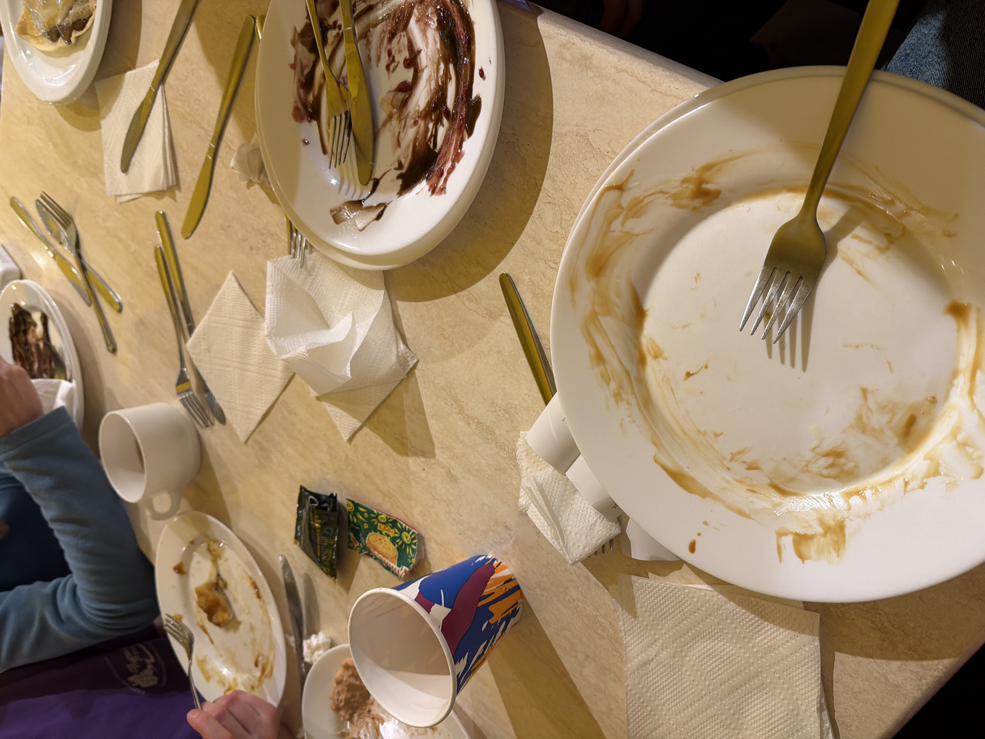 A table surface displays several dirty plates with leftover food scraps and condiments. Gold-colored forks and knives lie beside the plates, along with folded paper napkins. A white mug and a paper cup are also on the table, as well as individual packets of condiments. A person's arm and hand are visible, holding silverware near a plate. The setting appears to be indoors, likely at a restaurant or dining area. There's no indication this photo was taken in Berezniki, Russia.