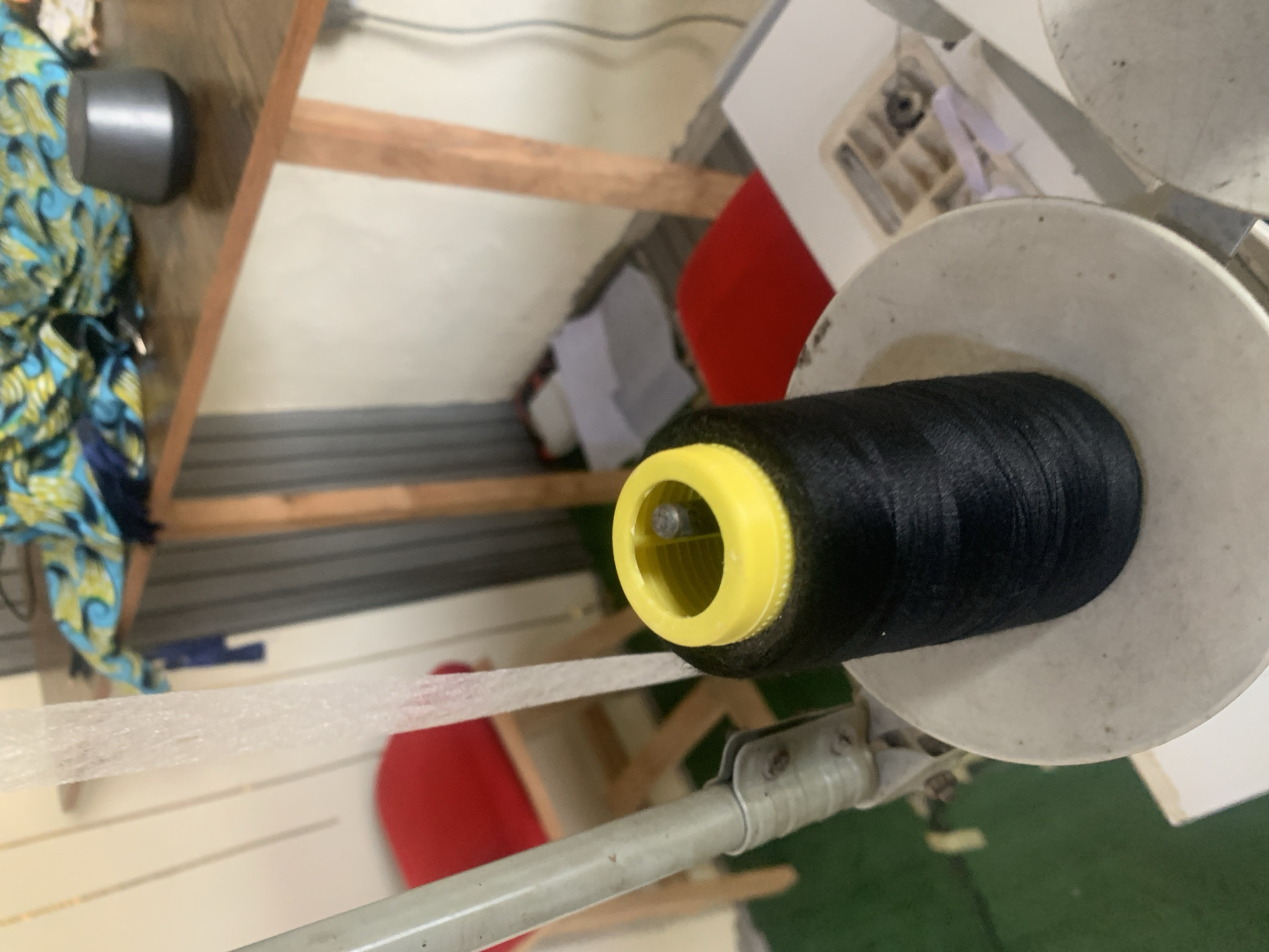 The image captures a close-up view of a black thread spool with a yellow top, positioned on a white base. A clear, thin plastic or elastic material drapes down from above, to the left of the spool. In the background, a wooden table holds a colorful fabric with a blue and green pattern and a small, dark, rounded object. Behind the table, a metal corrugated wall is visible. To the left, a portion of a red chair with a wooden frame can be seen. The scene appears to be an indoor setting, possibly a workshop or tailoring area, in the city of Jauro Yinu, Nigeria.